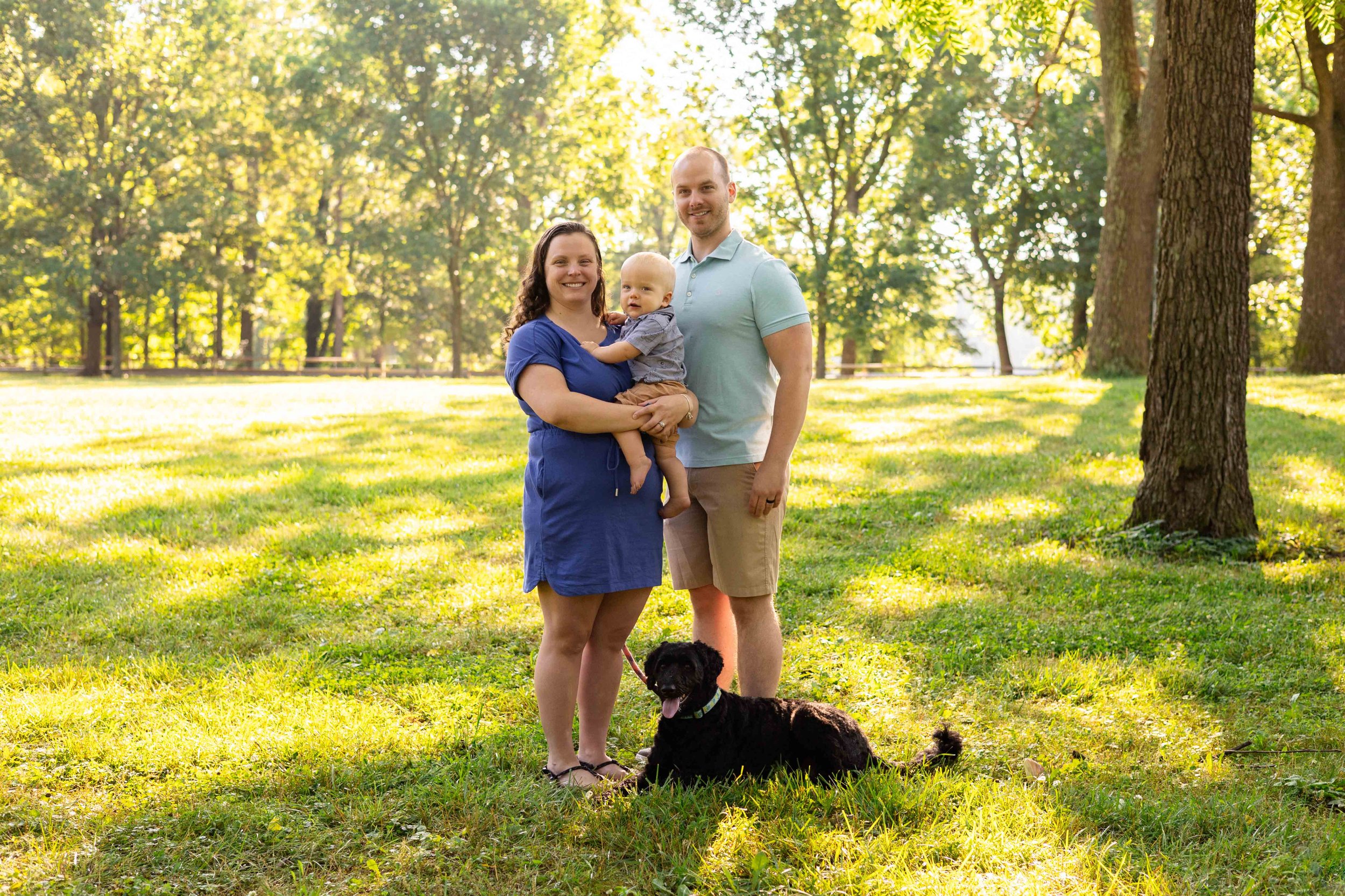A family with a baby and a dog stand under trees while wearing blue outfits - family photo taken by Jennifer Fulton Photography in Central NJ