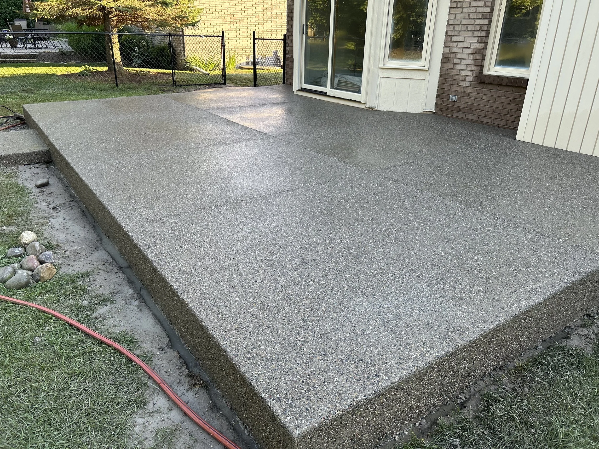 Newly poured concrete patio with textured surface next to house, surrounded by grass and a black fence.