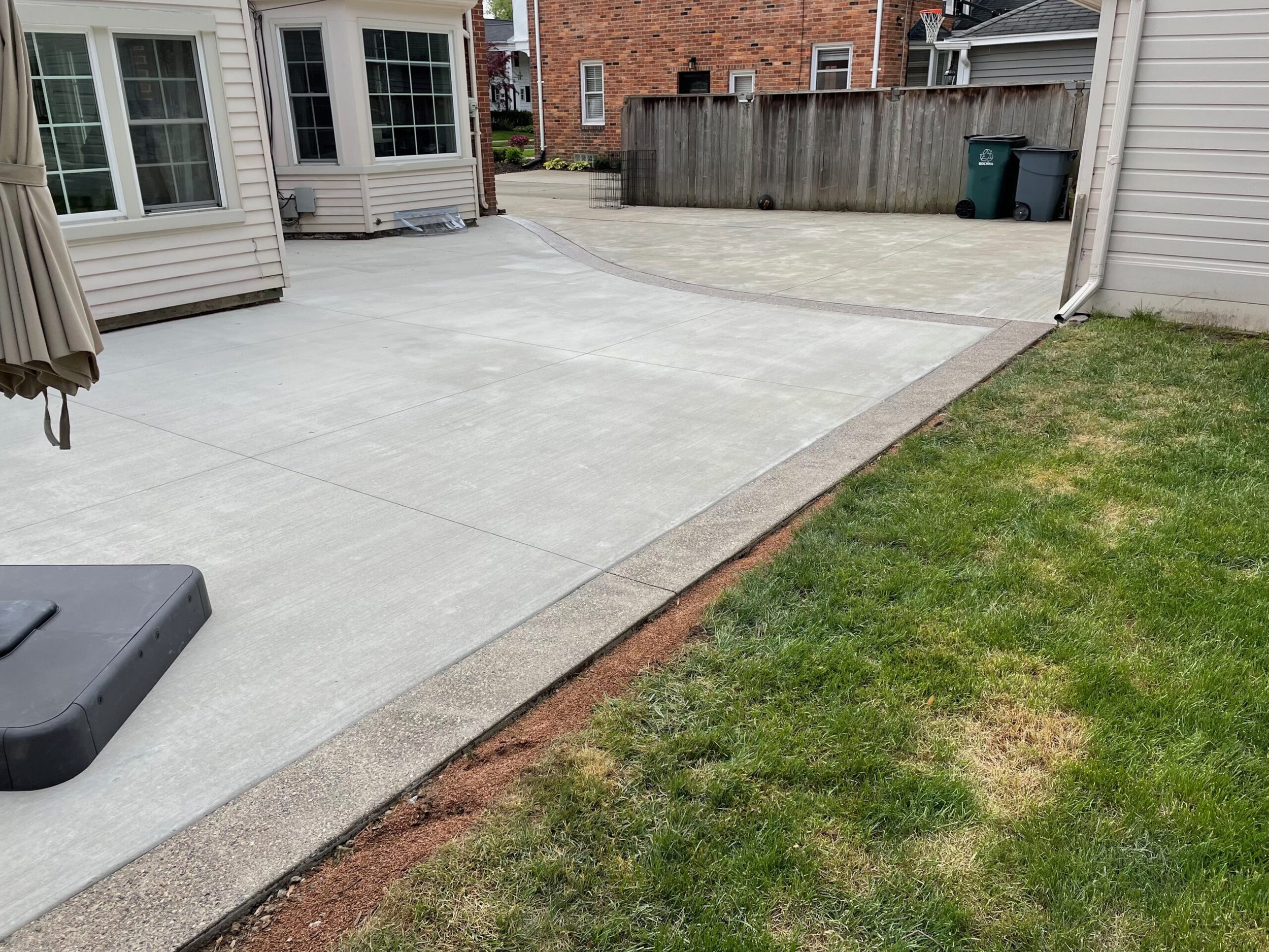 Newly paved concrete patio and driveway in a backyard with green grass, adjacent to a white house and a wooden fence, featuring trash bins and a basketball hoop.