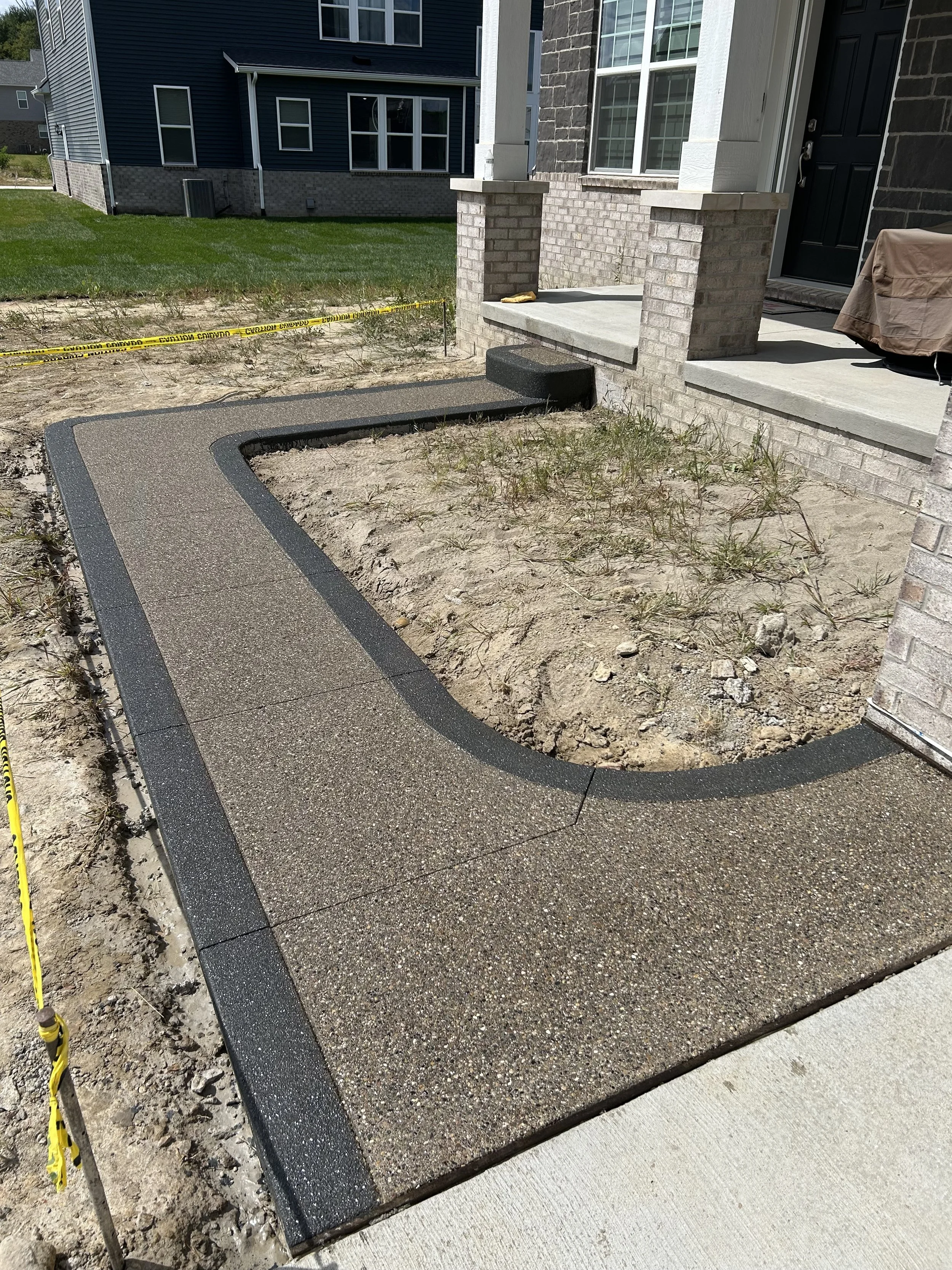A newly constructed concrete sidewalk with a curved section leading to a house entrance. The house has a brick exterior, and there is a small step with a black door. Construction caution tape is visible in the background.