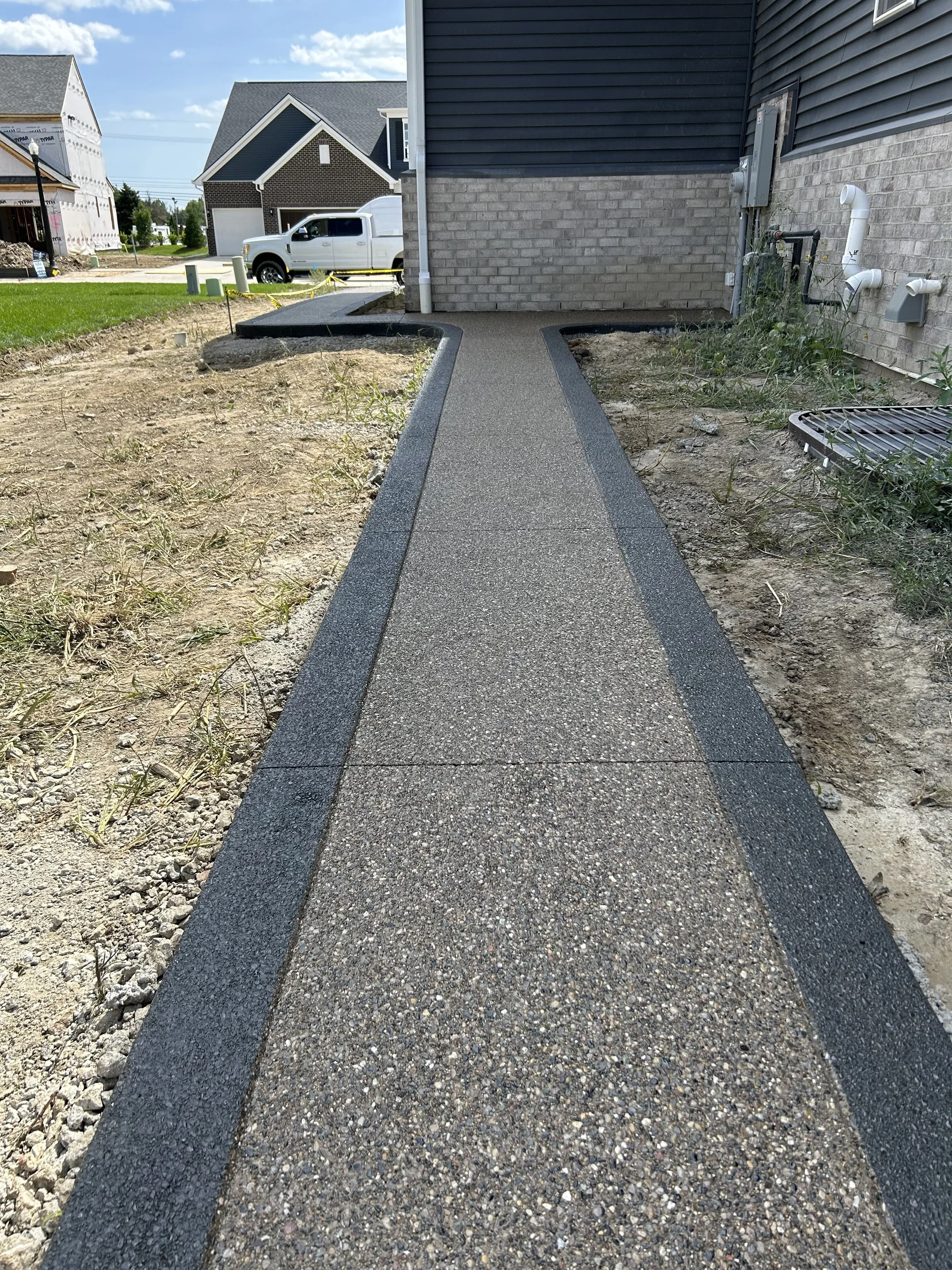 Concrete sidewalk path leading to a house, with a small area of dirt and grass on the sides, and neighboring houses in the background.