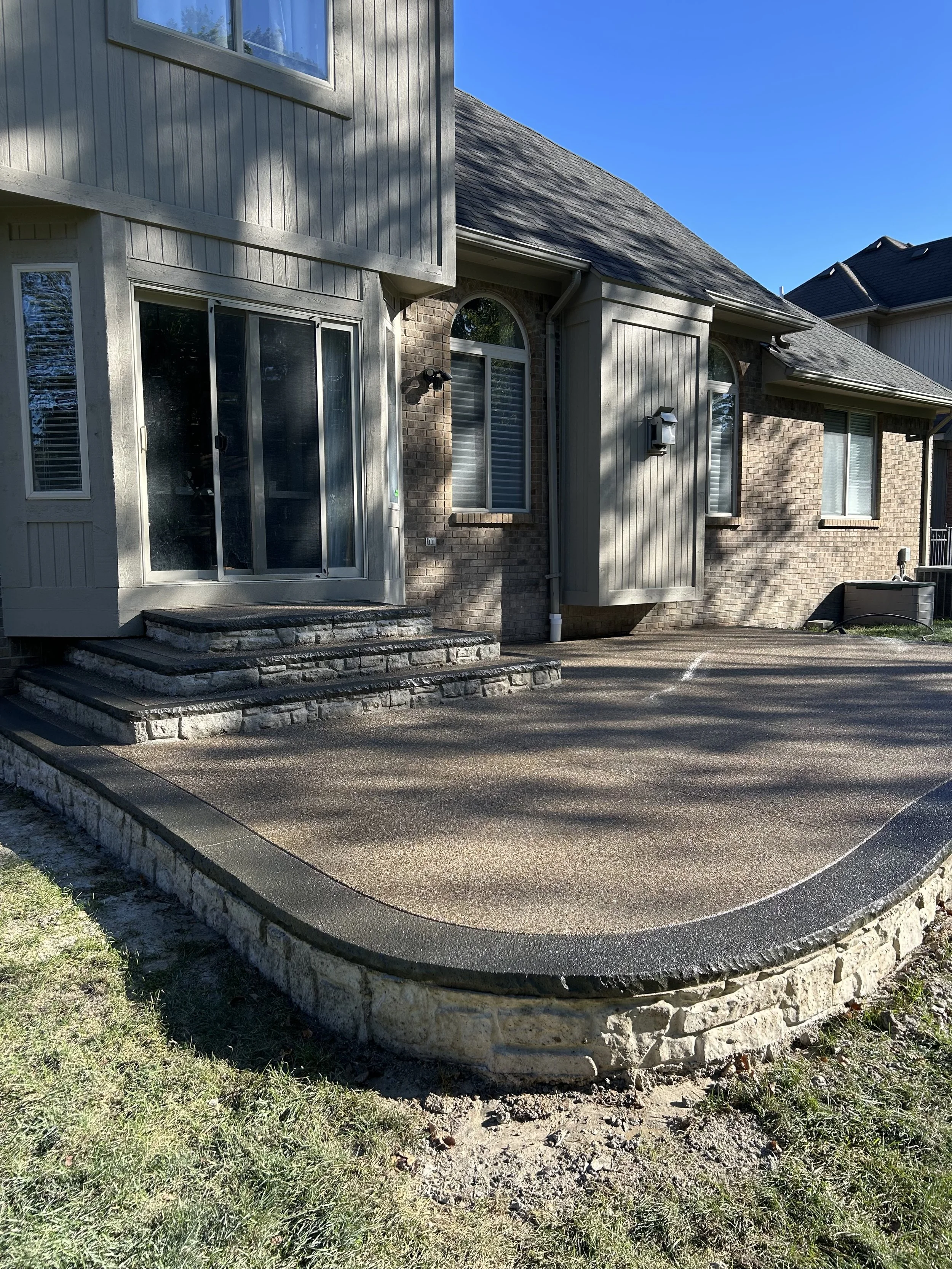 Back patio area with stone steps leading to a sliding glass door, brick and siding exterior house, and a concrete or textured surface patio with a curved edge, in a sunny outdoor setting.