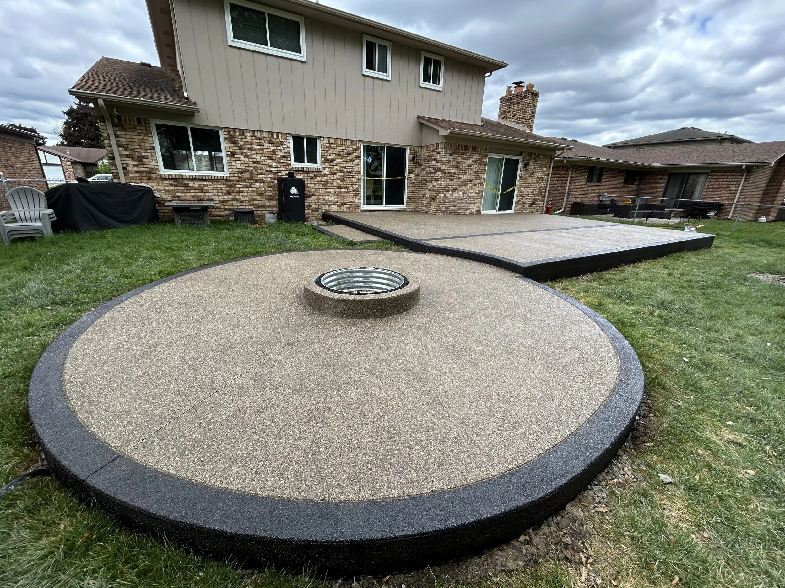 Backyard with a fire pit and a recently laid concrete deck, surrounded by grassy lawn, next to a brick house with sliding glass door and windows under a cloudy sky.