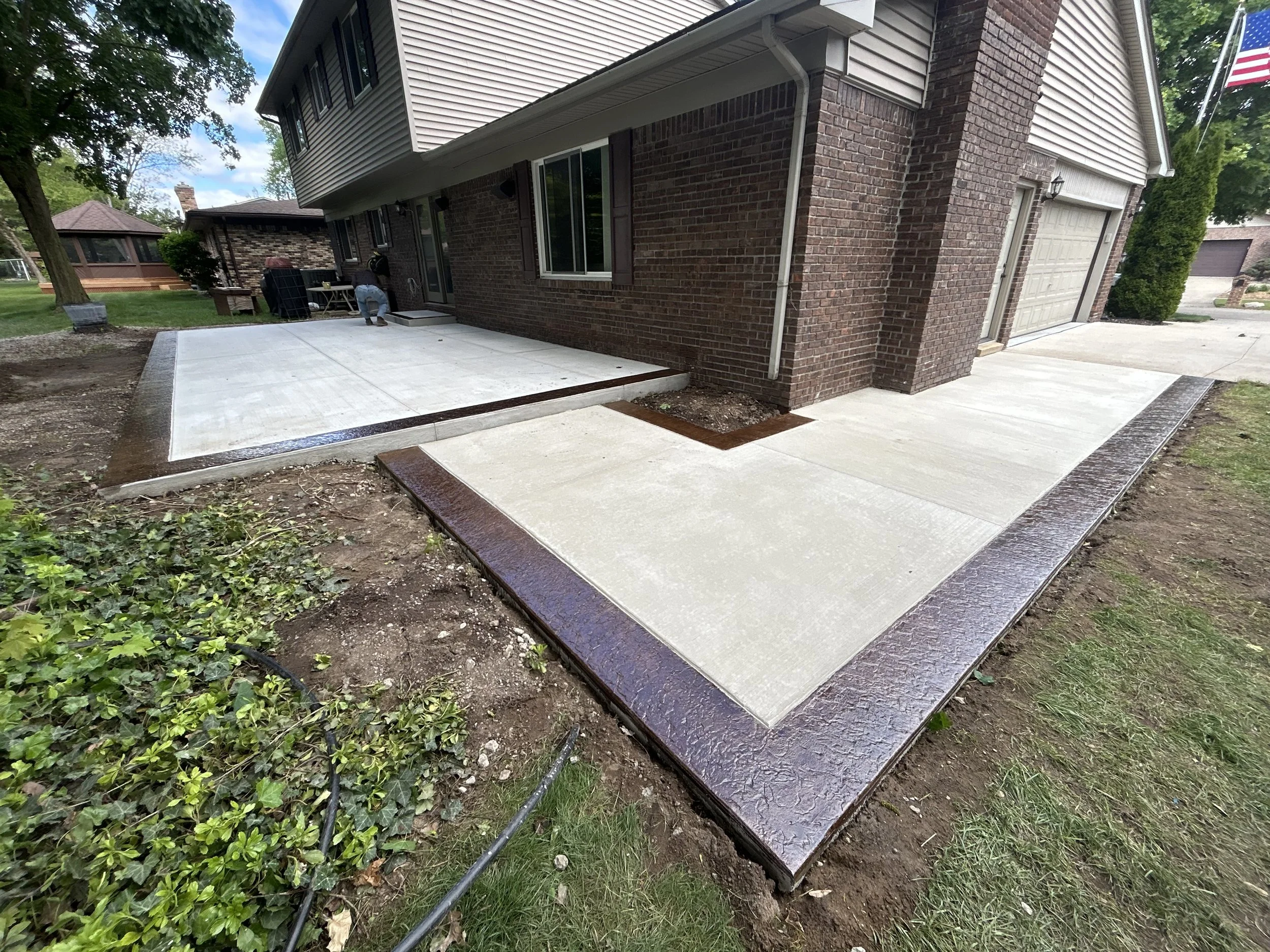 Newly constructed concrete patio with a decorative brown border surrounding it, attached to the back of a brick house, with a man working on finishing touches. The area is partly landscaped with some grass and bushes.