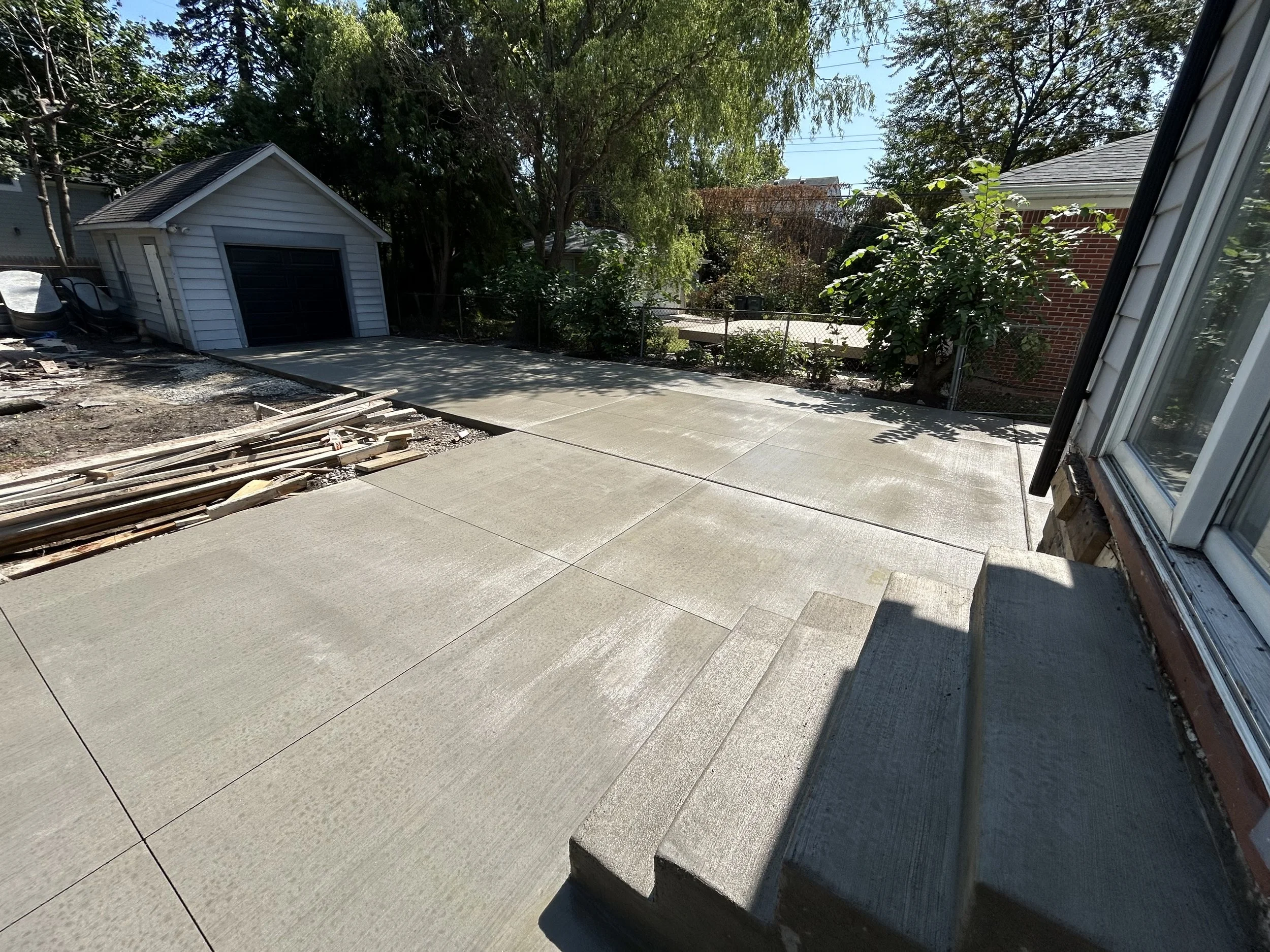 View of a backyard with a newly poured concrete patio, a small white shed, and some gardening tools and materials on the left side. There are trees and a fenced garden area with plants and bushes in the background, and the house's exterior wall with