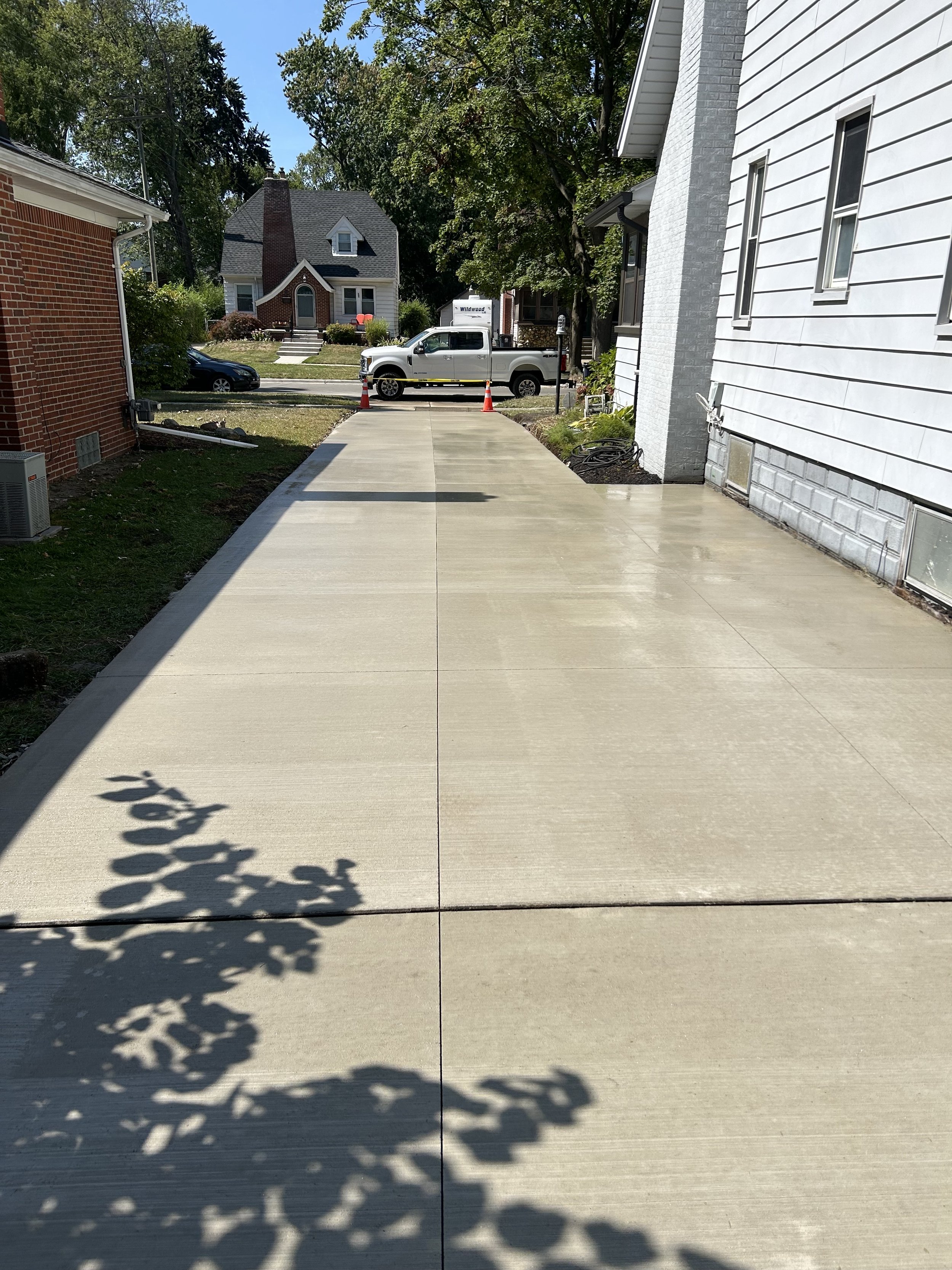 Concrete driveway in a residential neighborhood with houses, trees, and parked cars; orange traffic cones at the end of the driveway.