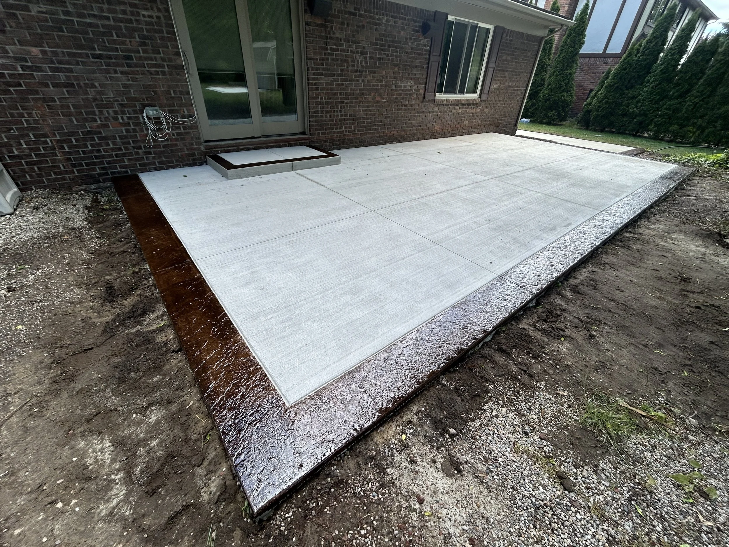 Newly poured concrete patio at the back of a brick house with surrounding dirt and gravel.