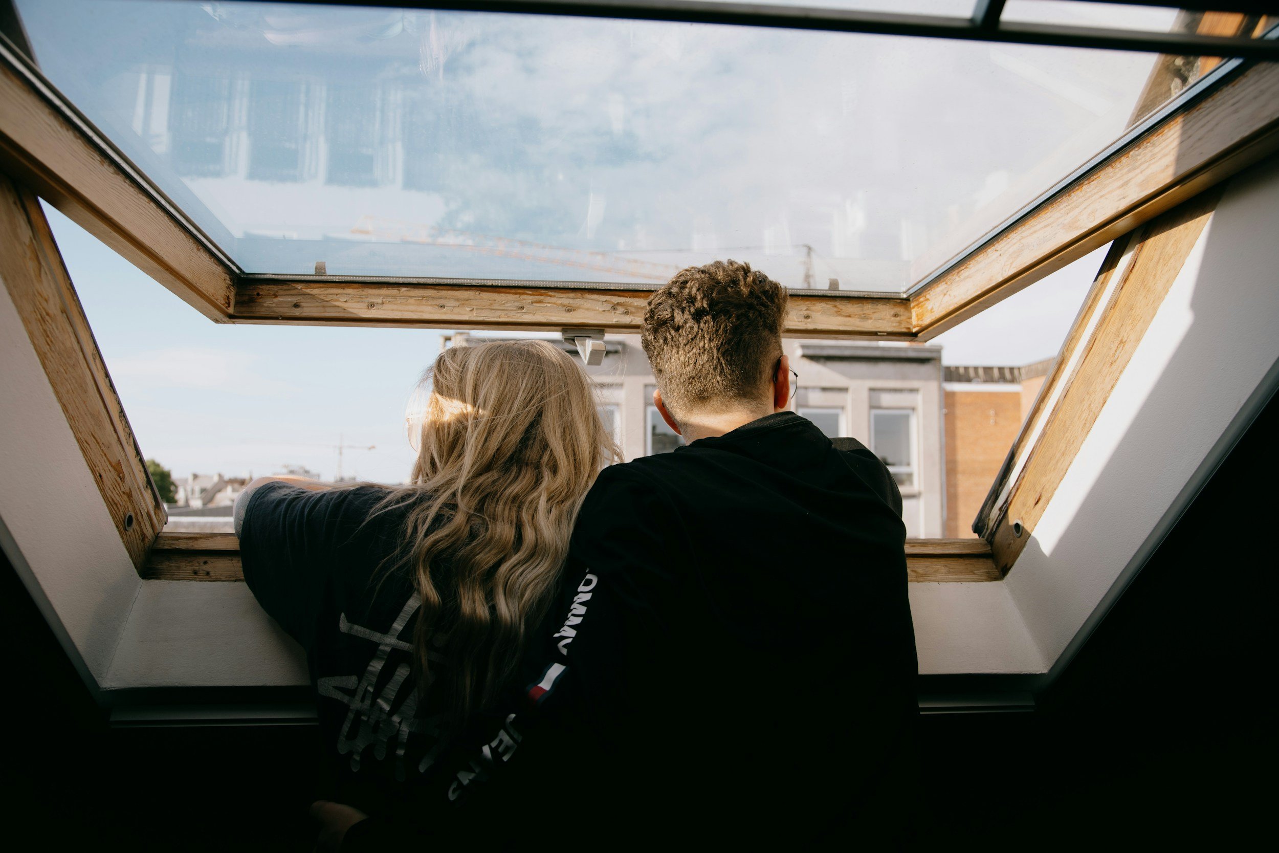 Two people are looking out of an open skylight window onto a cityscape with buildings and a clear sky.