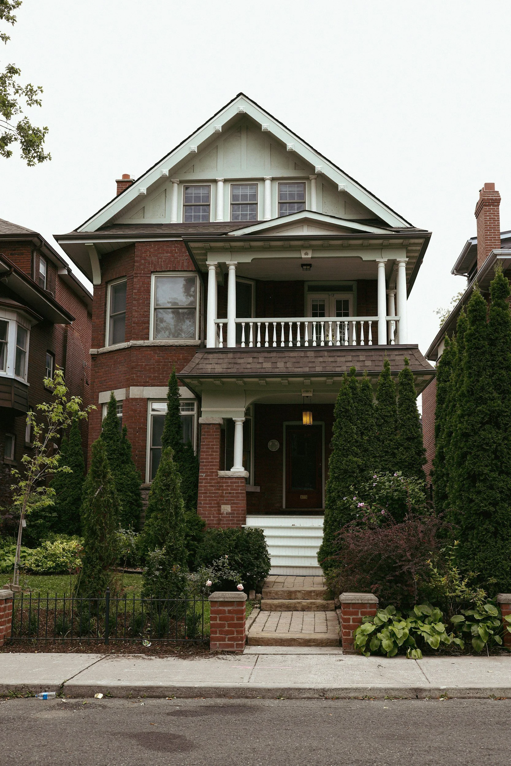 A multi-story brick house with a porch and balcony, surrounded by tall green trees and shrubs, with a brick walkway leading to the front steps.