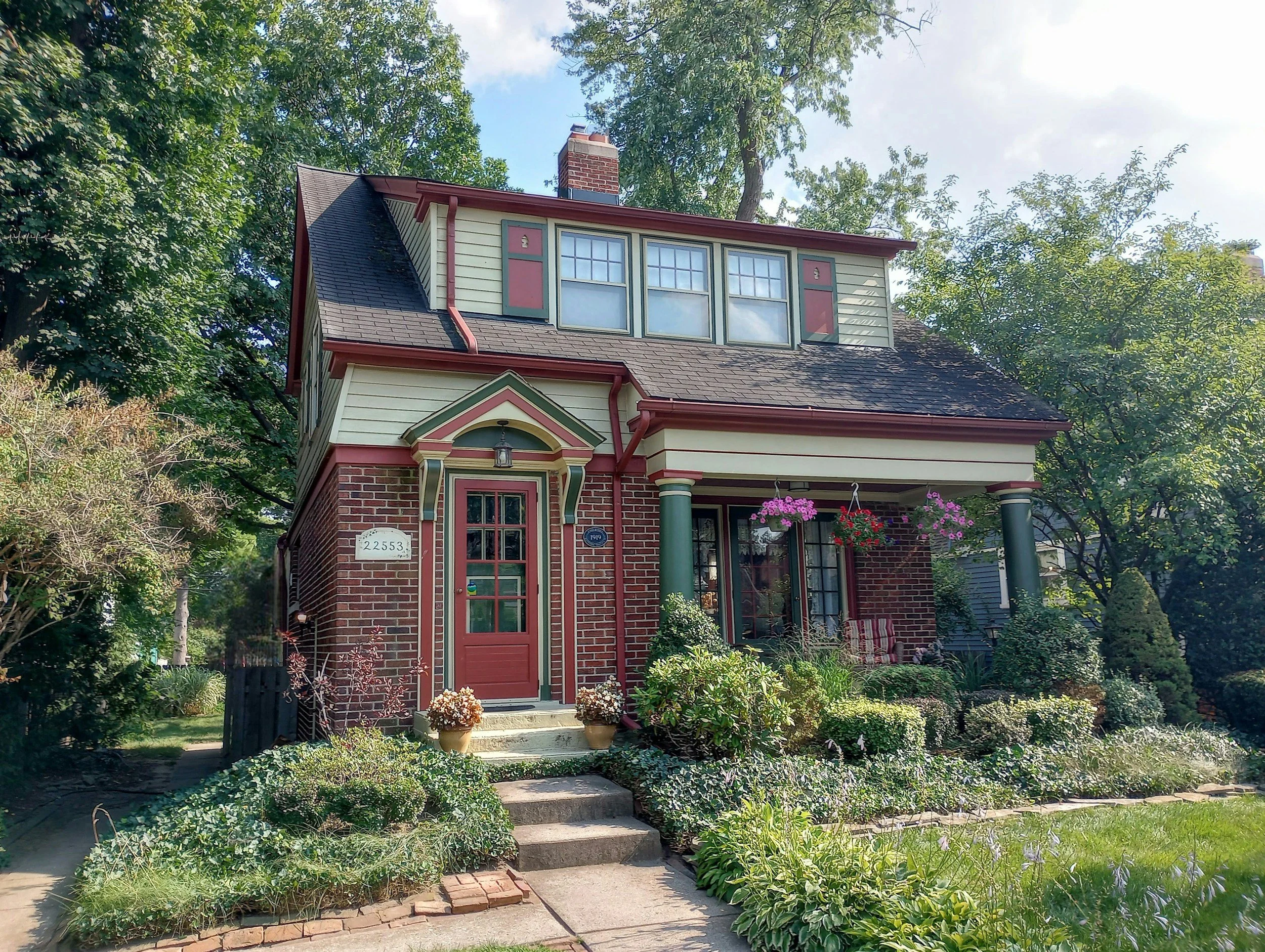 A charming two-story house with a brick and wood exterior, featuring a front porch with hanging flower baskets, surrounded by lush garden plants, and tall trees in the background under a partly cloudy sky.