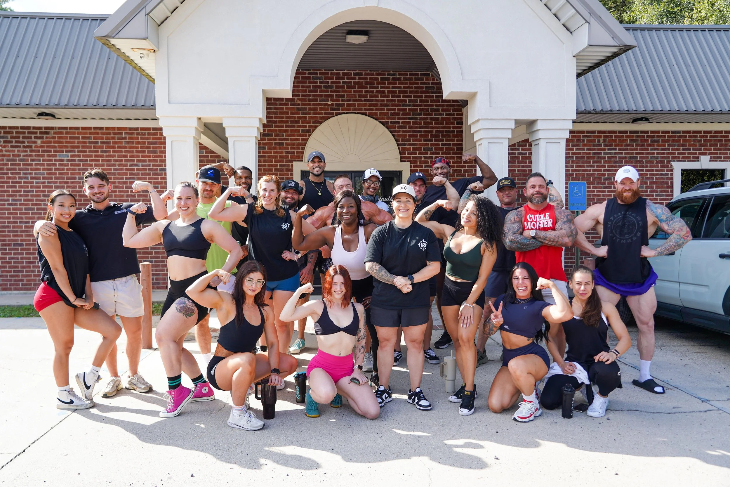 Group of diverse fitness people posing outside in front of a brick building, flexing their muscles and smiling.