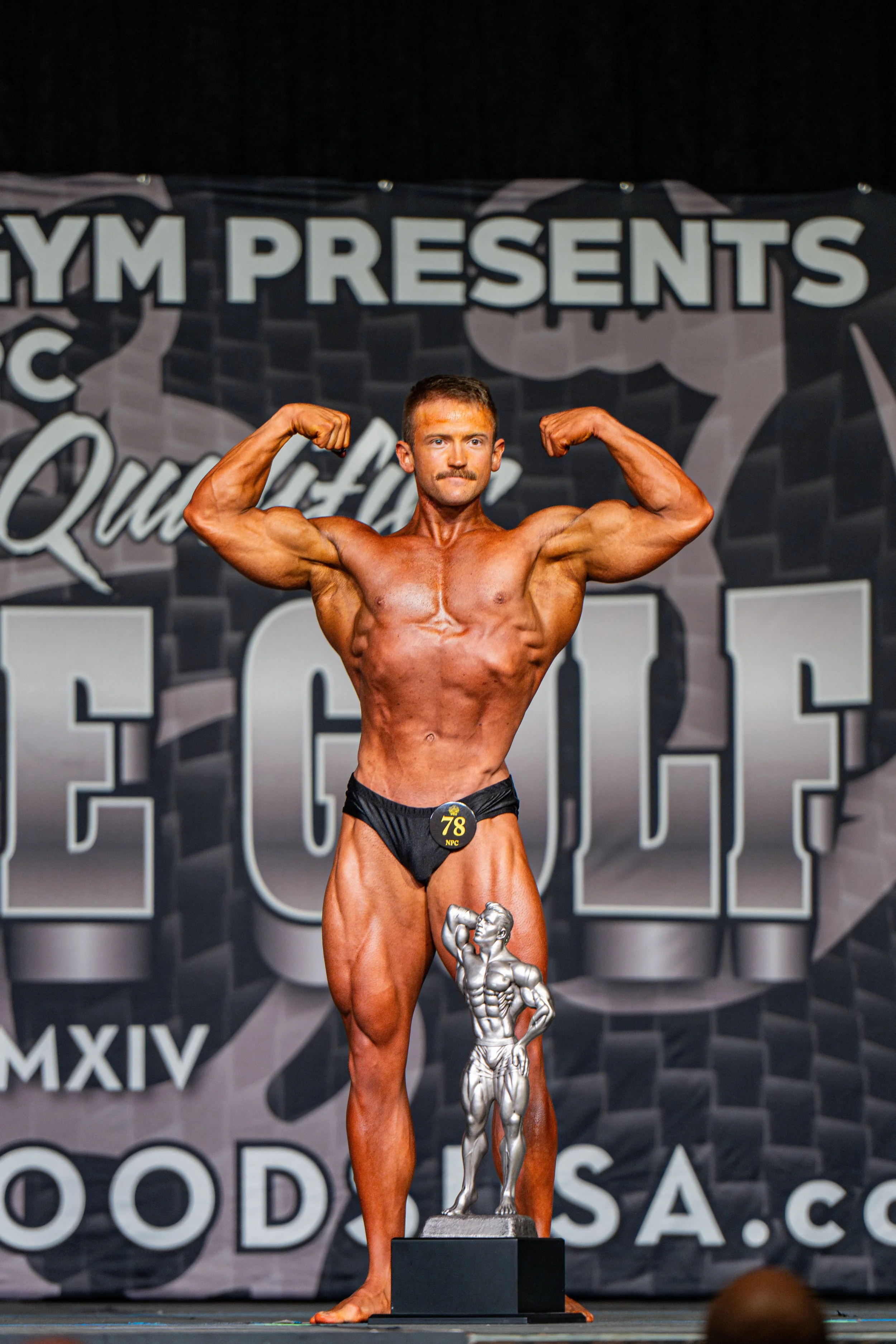 Male bodybuilder on stage, flexing muscles in a bodybuilding competition, wearing black briefs, with a trophy at his feet, and a backdrop with event branding.