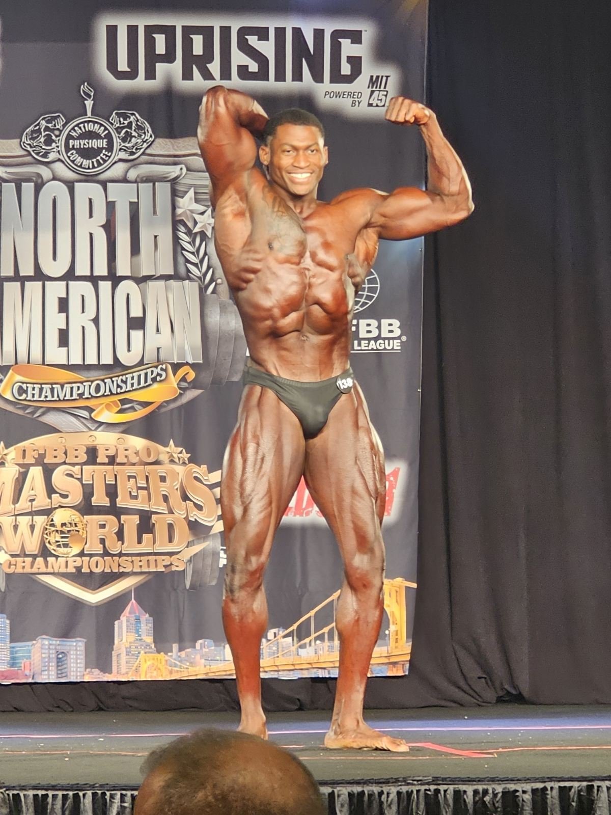 A male bodybuilder on stage at a bodybuilding competition, flexing his muscles and smiling. The background features banners indicating the event is the North American Championships and the IFBB Pro Masters World Championships.