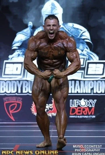 Bodybuilder posing on stage during a competition, flexing muscles, with a championship backdrop featuring logos and the year 2023.