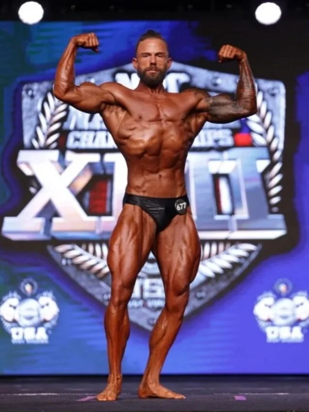 Bodybuilder flexing muscles on stage with Pro Tan and NPC News Online logos.