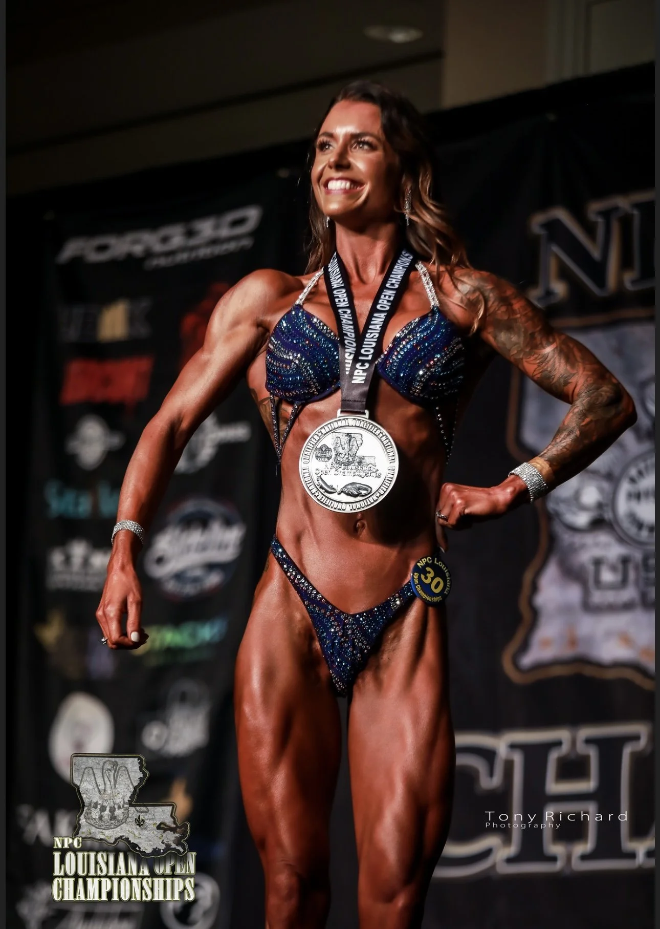 A female bodybuilder posing on stage at the NPC Louisiana Open Championships, wearing a sparkling bikini and a medal around her neck.