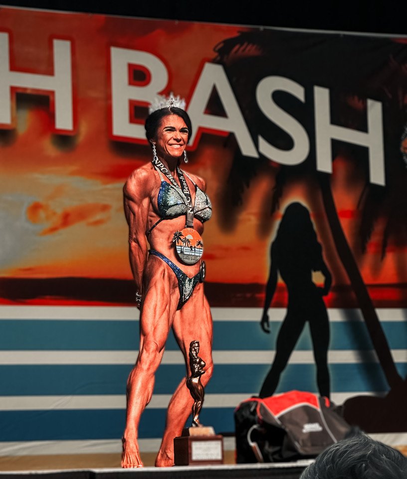 Female bodybuilder on stage wearing a medal and holding a trophy, posing in a bikini with a backdrop reading "BASH."