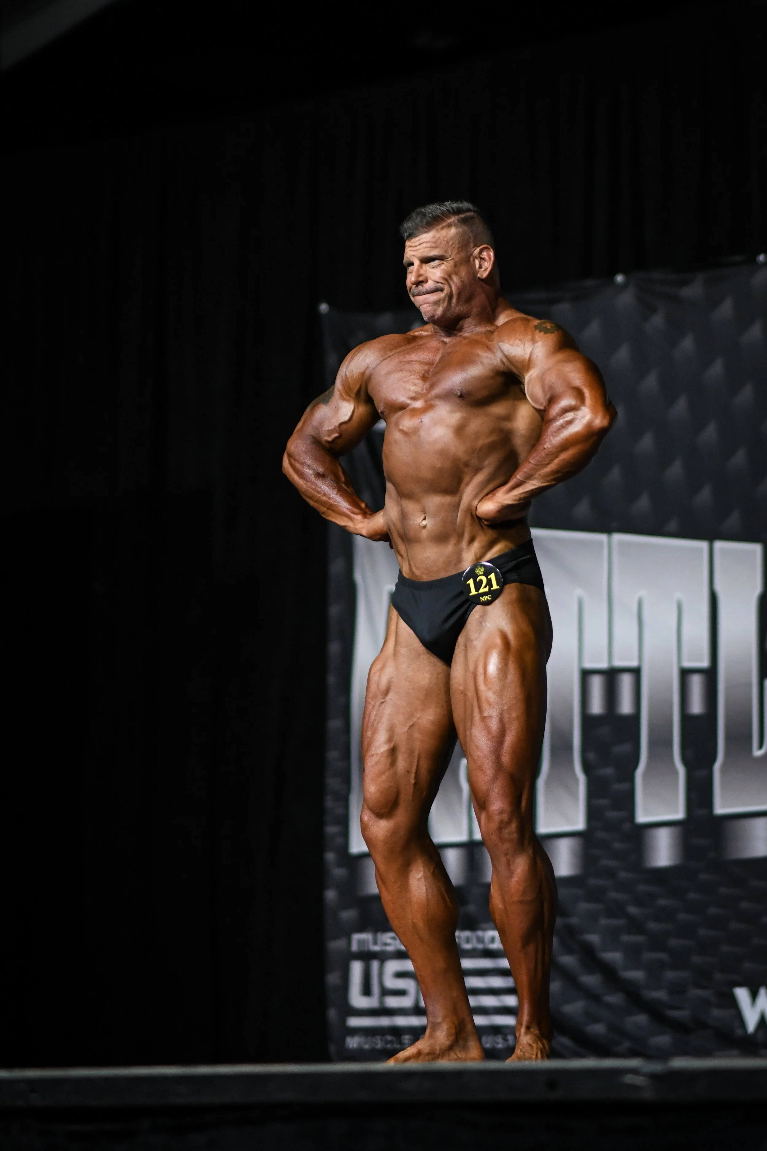 A male bodybuilder in black posing trunks standing on stage with hands on hips, showcasing muscular physique at a bodybuilding competition.
