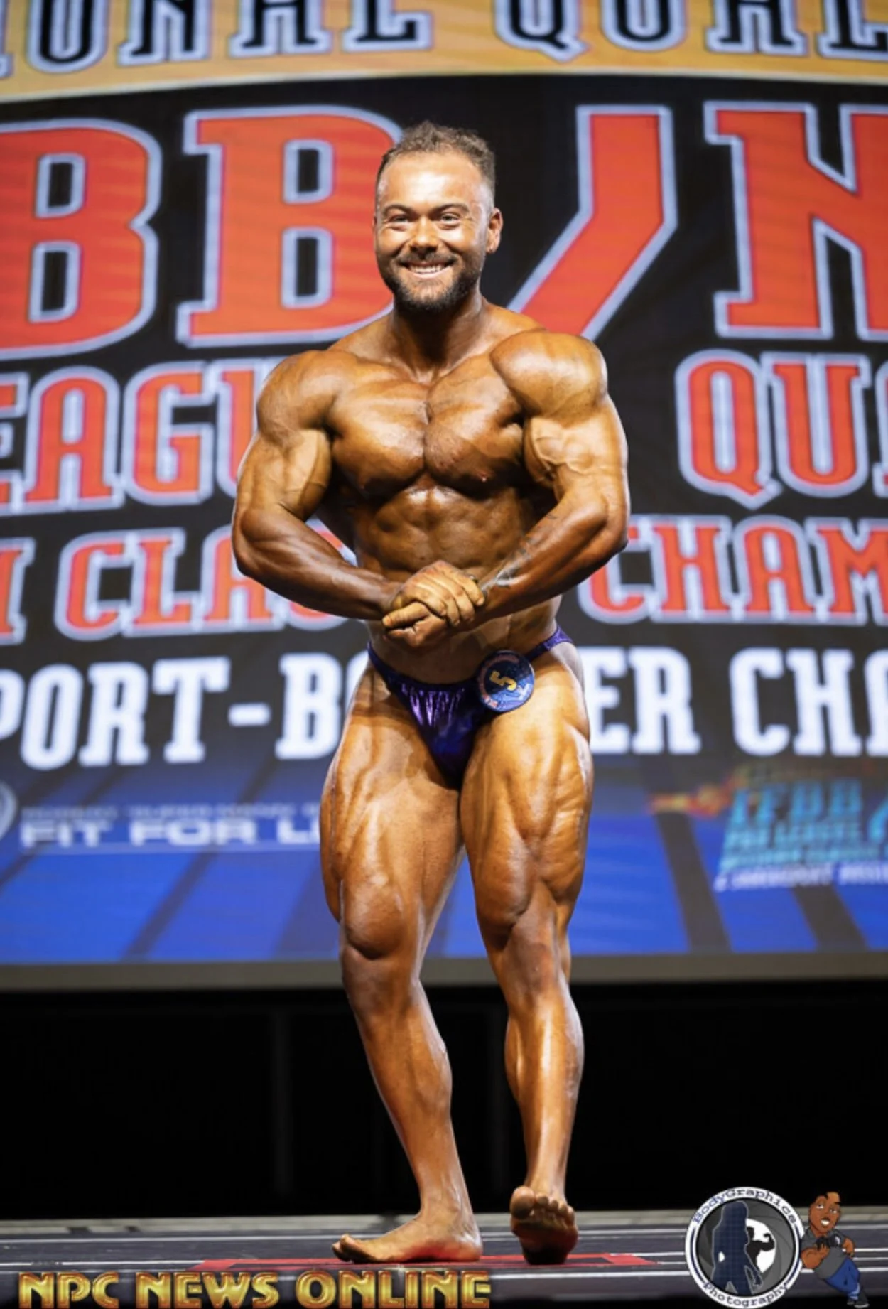 A male bodybuilder with tan skin and a beard on stage during a bodybuilding competition, wearing a purple posing suit with the number 5, smiling and flexing his muscles. The background displays the event name and sponsors.