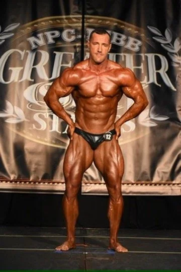 Male bodybuilder posing on stage during a competition, with a muscular physique and tanned skin, standing in front of a backdrop with "NPC" logo and text.