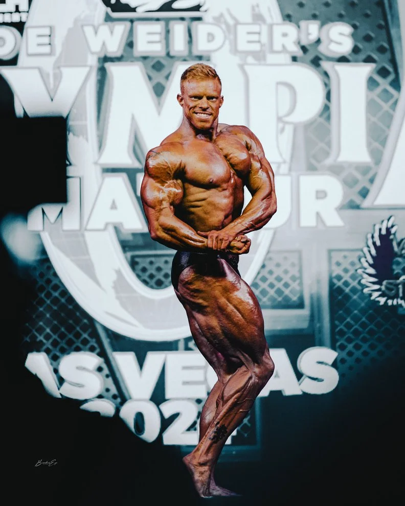 Bodybuilder posing on stage at a competition, showing muscular physique against a backdrop with text "Olympia" and "Las Vegas."