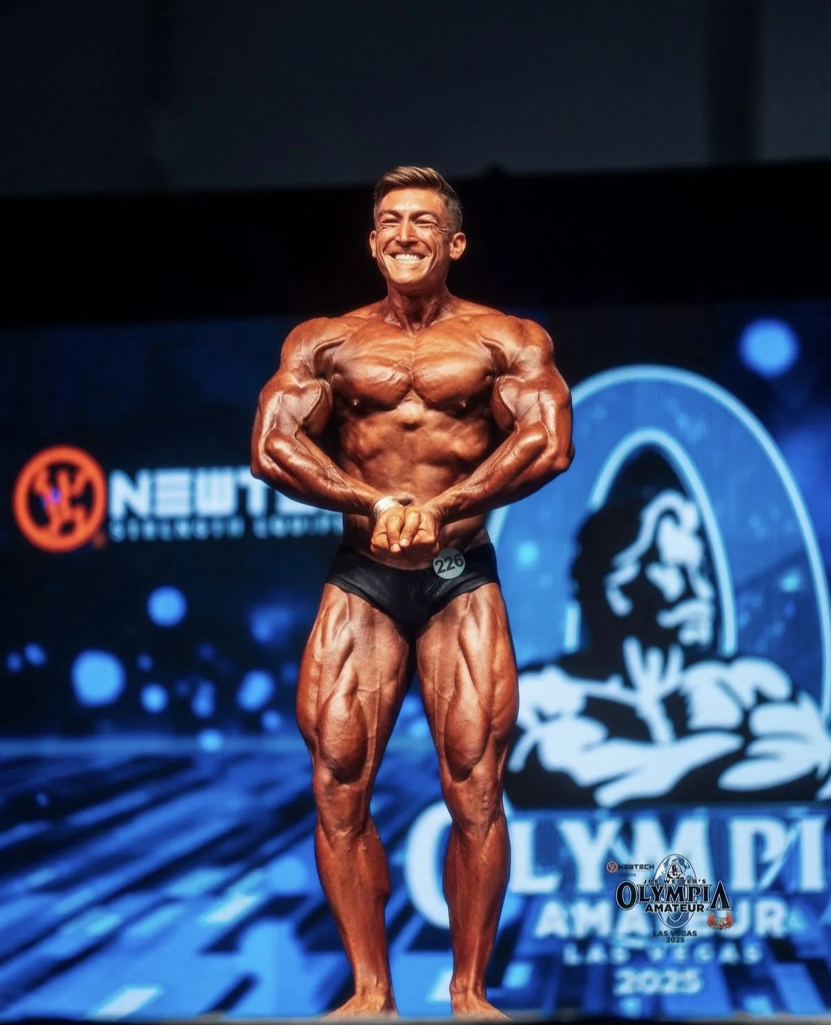 A male bodybuilder on stage during a bodybuilding competition, flexing his muscles and smiling while wearing black shorts, with a backdrop displaying text and logos related to the event.
