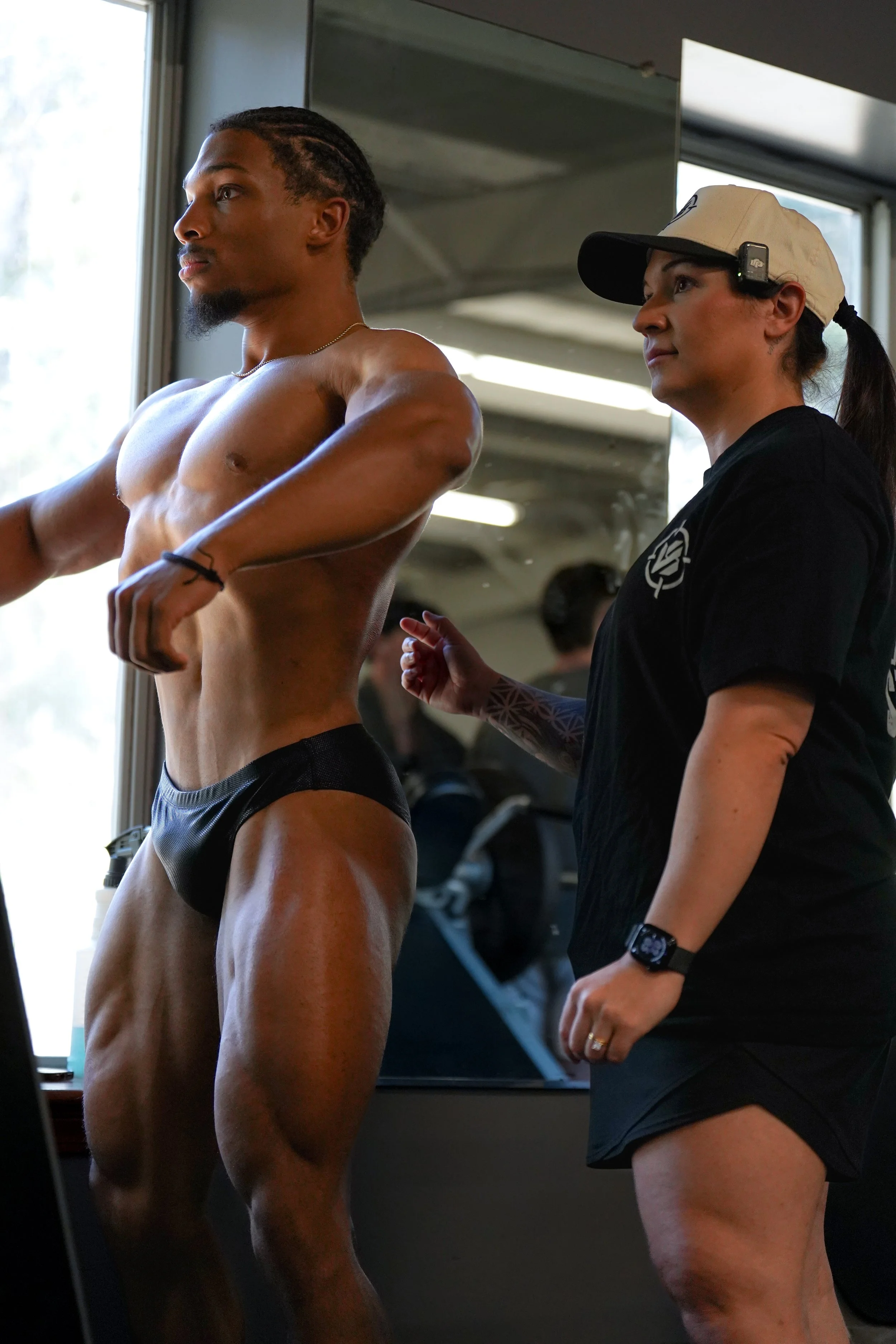 A shirtless man with muscular build and dark skin standing in a gym, being assisted by a woman wearing a beige cap, black t-shirt, and a fitness tracker on her wrist.