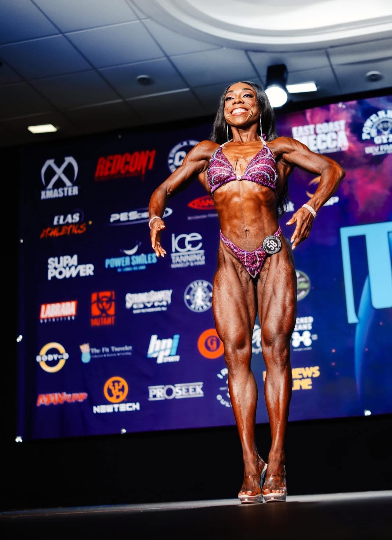 A female bodybuilder in a purple bikini flexing on stage during a competition.