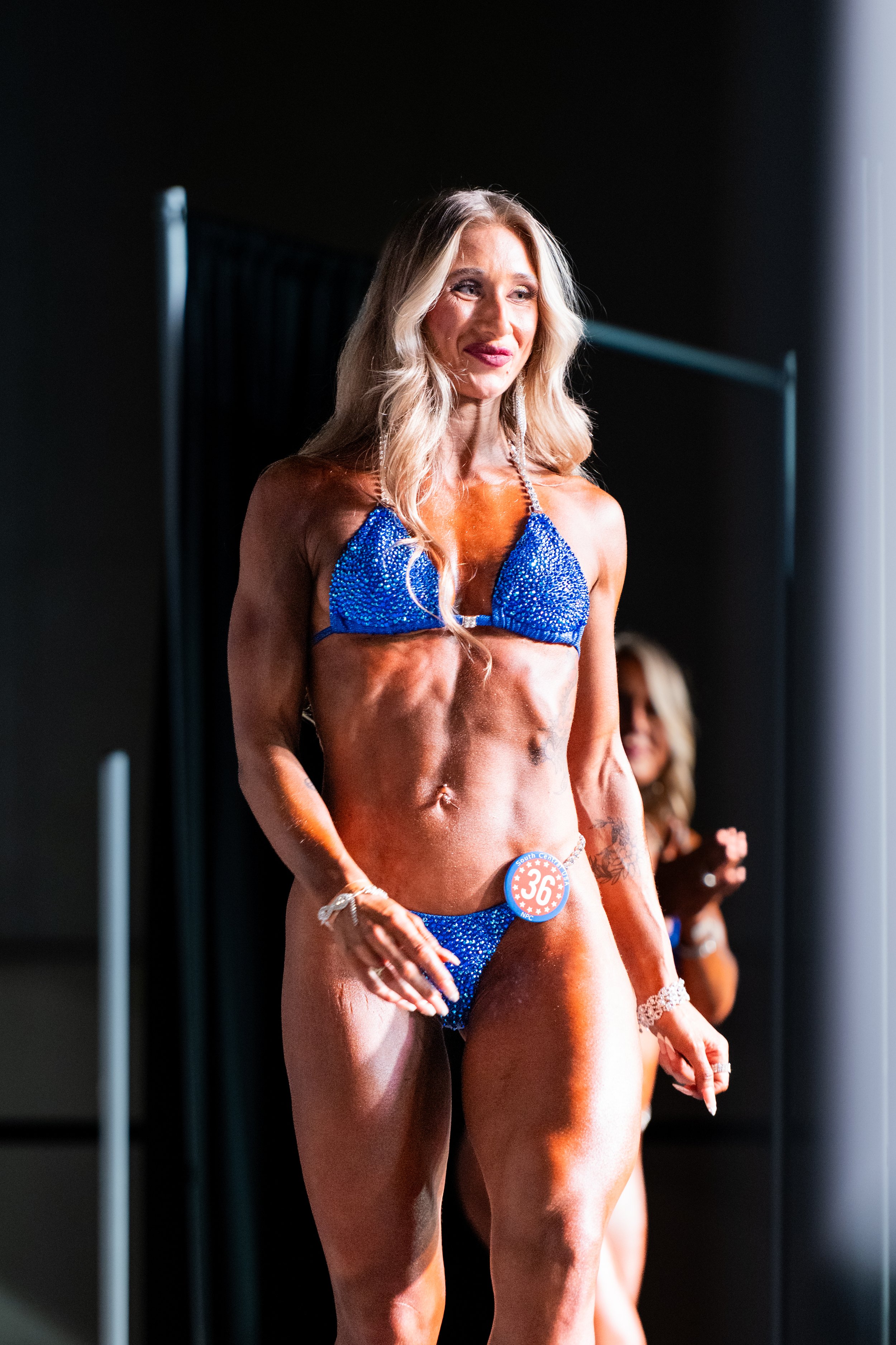 A woman in a blue bikini participating in a fitness or bodybuilding competition, wearing a contest number 36, with a blurred person in the background.