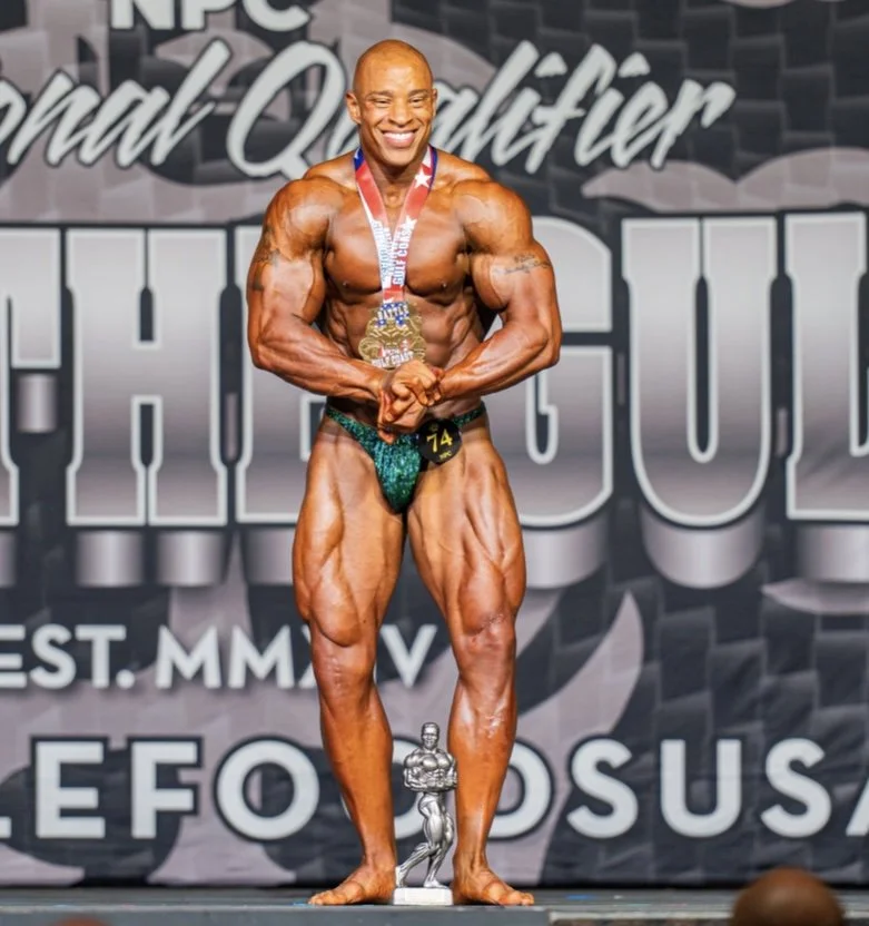 Male bodybuilder on stage at a fitness competition, smiling, wearing a medal around his neck, posing with flexed muscles, trophy on the floor in front, backdrop with event name.