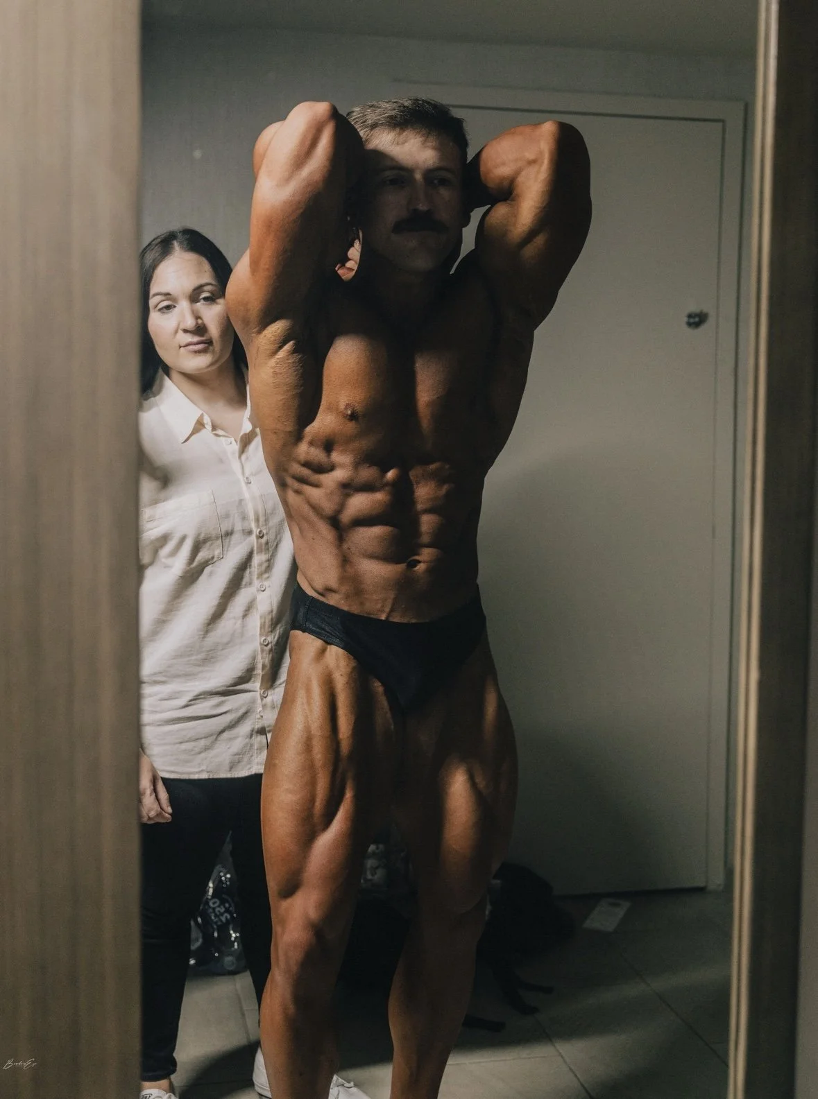 A muscular man in black posing underwear taking a mirror selfie in a room, with a woman in a white button-up shirt standing behind him and observing.