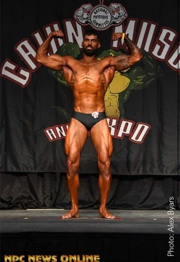 Bodybuilder posing on stage with "Cajun Muscle & Expo" banner.