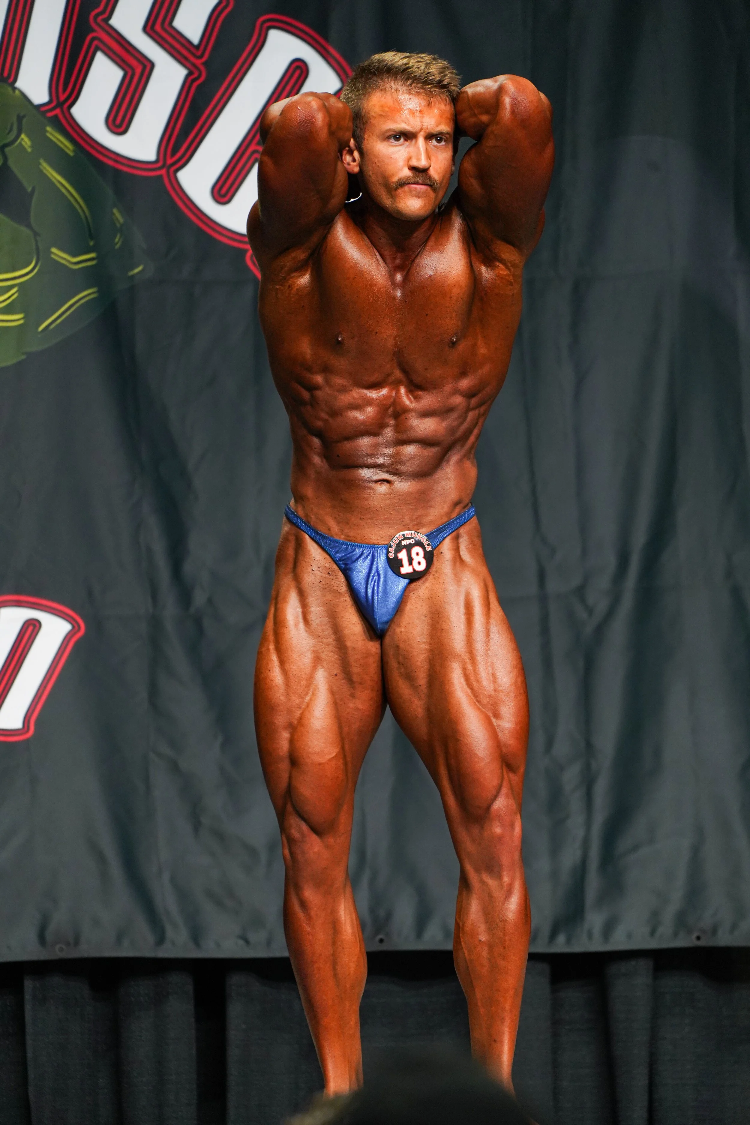 A male bodybuilder with tanned skin and muscular physique posing on stage during a competition, wearing a blue posing thong and a competition badge showing the number 18.