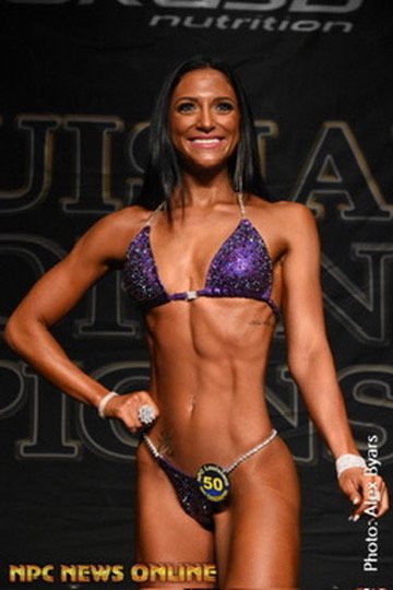 A female bodybuilder in a purple bikini poses on stage, showcasing her muscular physique. She is wearing a competition number 50 badge and has long dark hair. The background displays a competition banner.