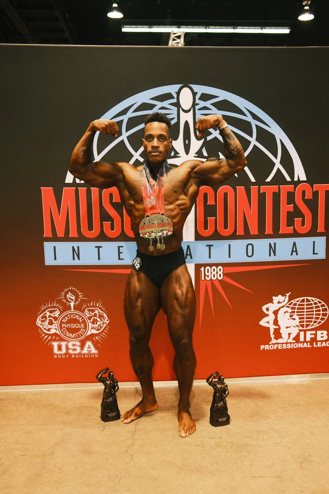 A male bodybuilder in black shorts flexing his biceps on stage at a bodybuilding competition, wearing medals around his neck and two trophies on the floor in front of him, with a backdrop displaying the words 'Muscle Contest International' and logos 
