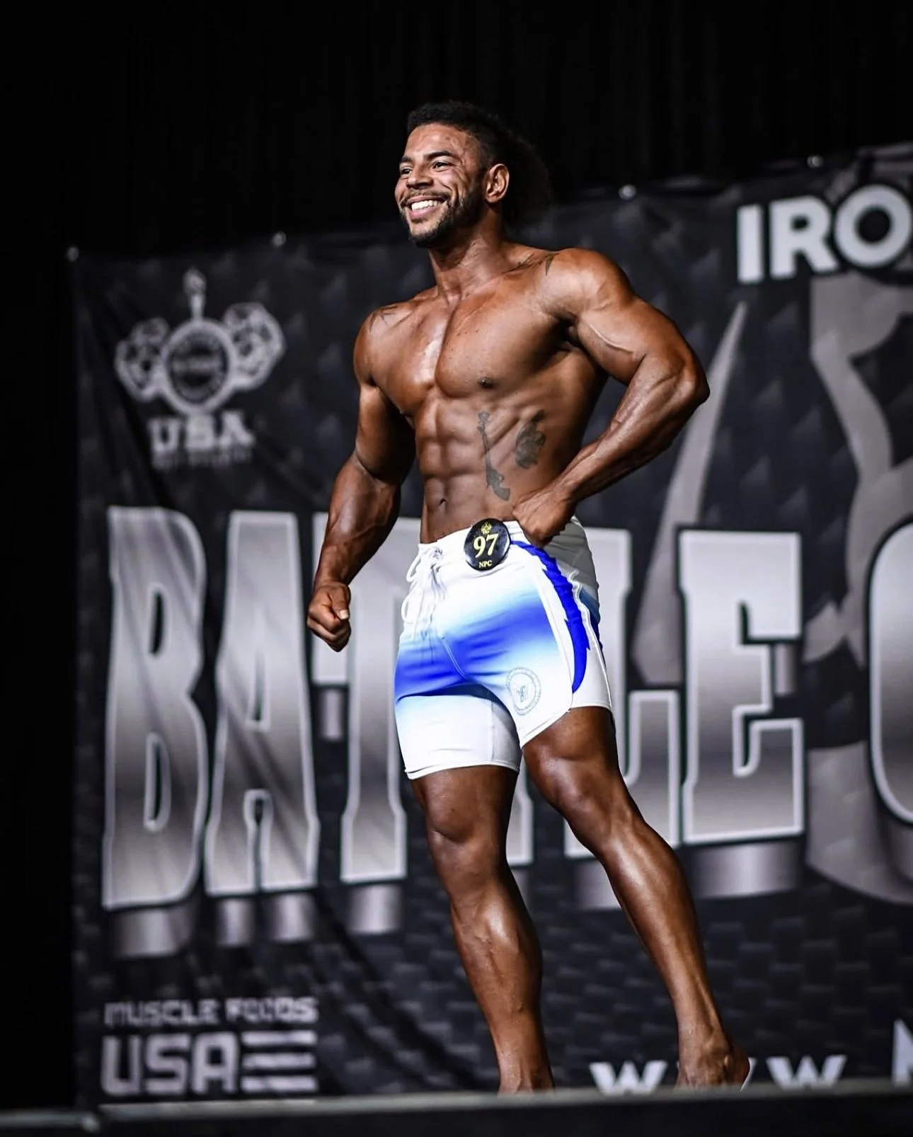 A male bodybuilding competitor standing on stage in a flexed pose, wearing white and blue shorts, smiling with a backdrop that reads 'IRON' and 'USA'.