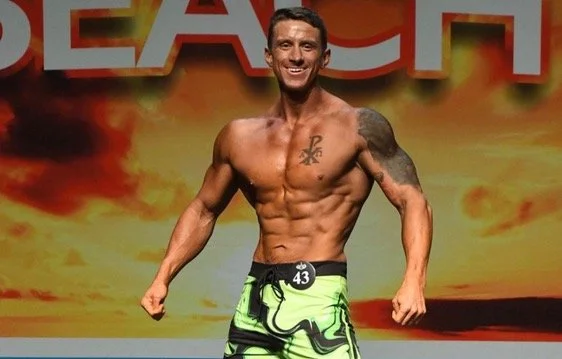 Bodybuilder poses confidently on stage wearing green shorts with number 43, muscles flexed.