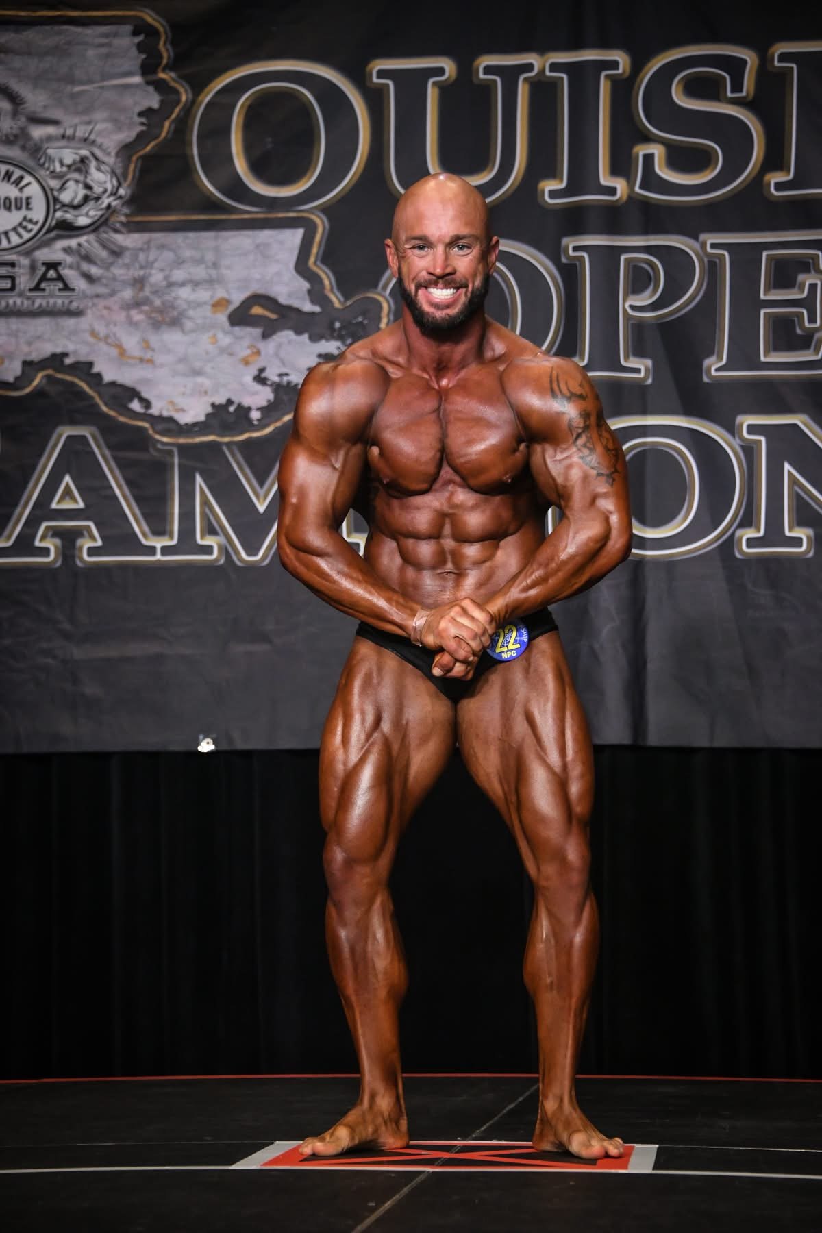 A muscular male bodybuilder with a shaved head, beard, and tan skin, posing on stage at a bodybuilding competition, flexing his biceps and showing his abs and leg muscles, with a backdrop that reads 'Outs' and 'Hope' in large letters.
