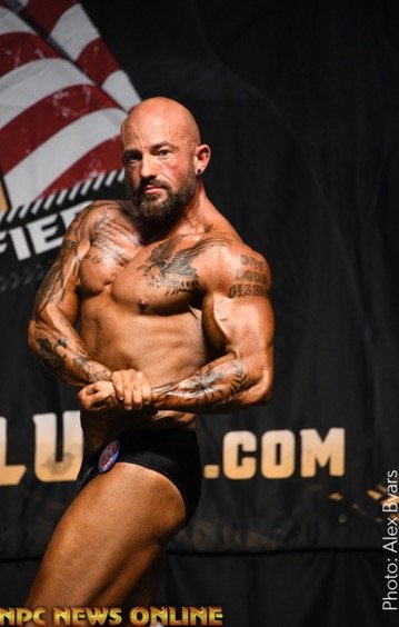 Bodybuilder posing on stage with tattoos, wearing black trunks, against a backdrop with American flag and logos.
