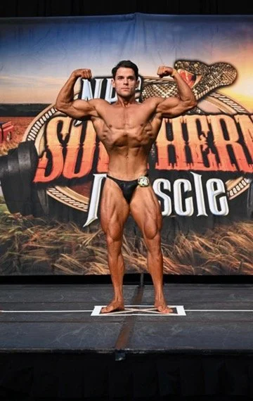 Bodybuilder posing on stage at a competition with a banner in the background.
