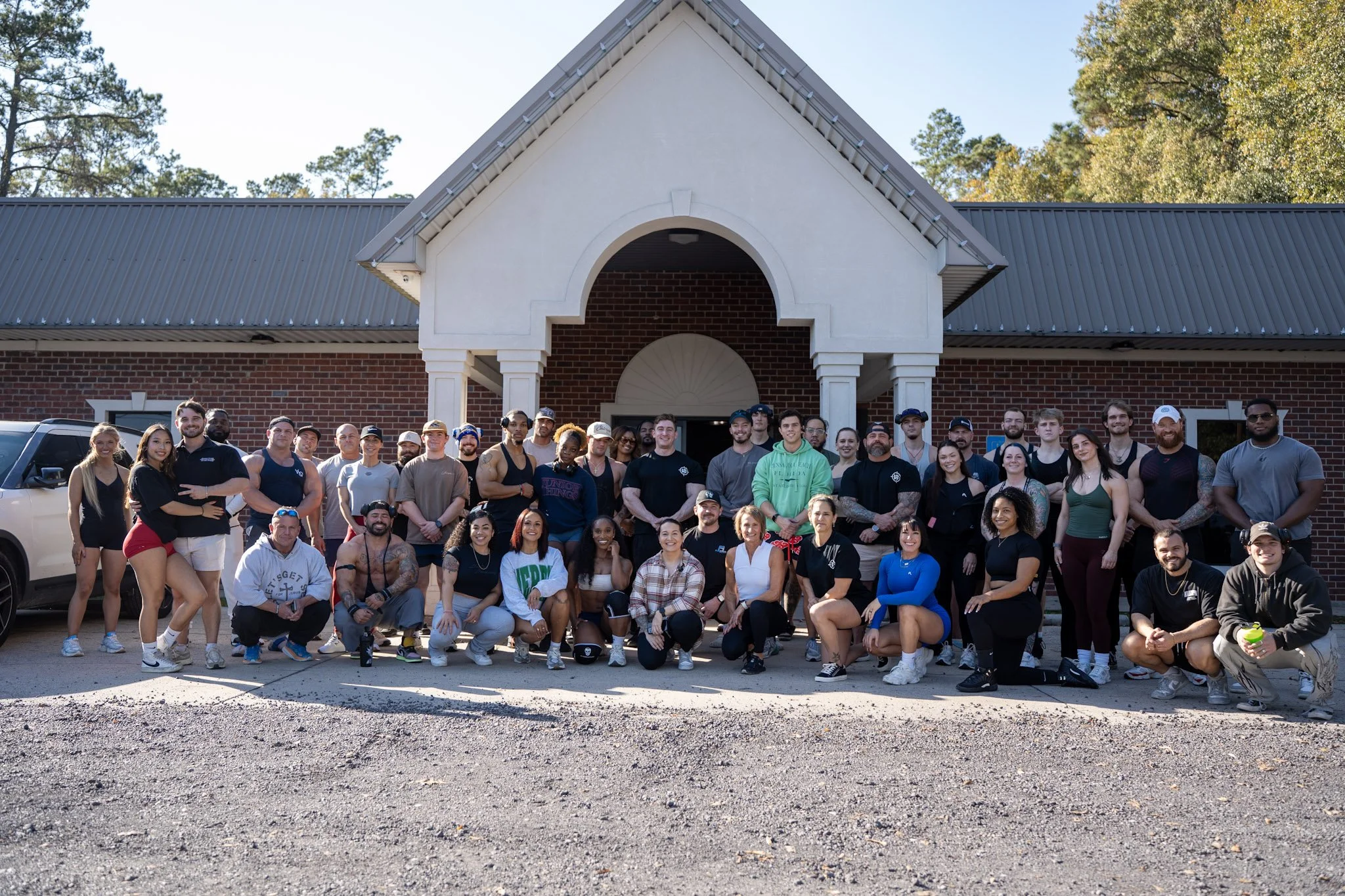 Large group of diverse people posing outside in front of a brick building with white accents and a gabled roof, trees in the background, on a sunny day.