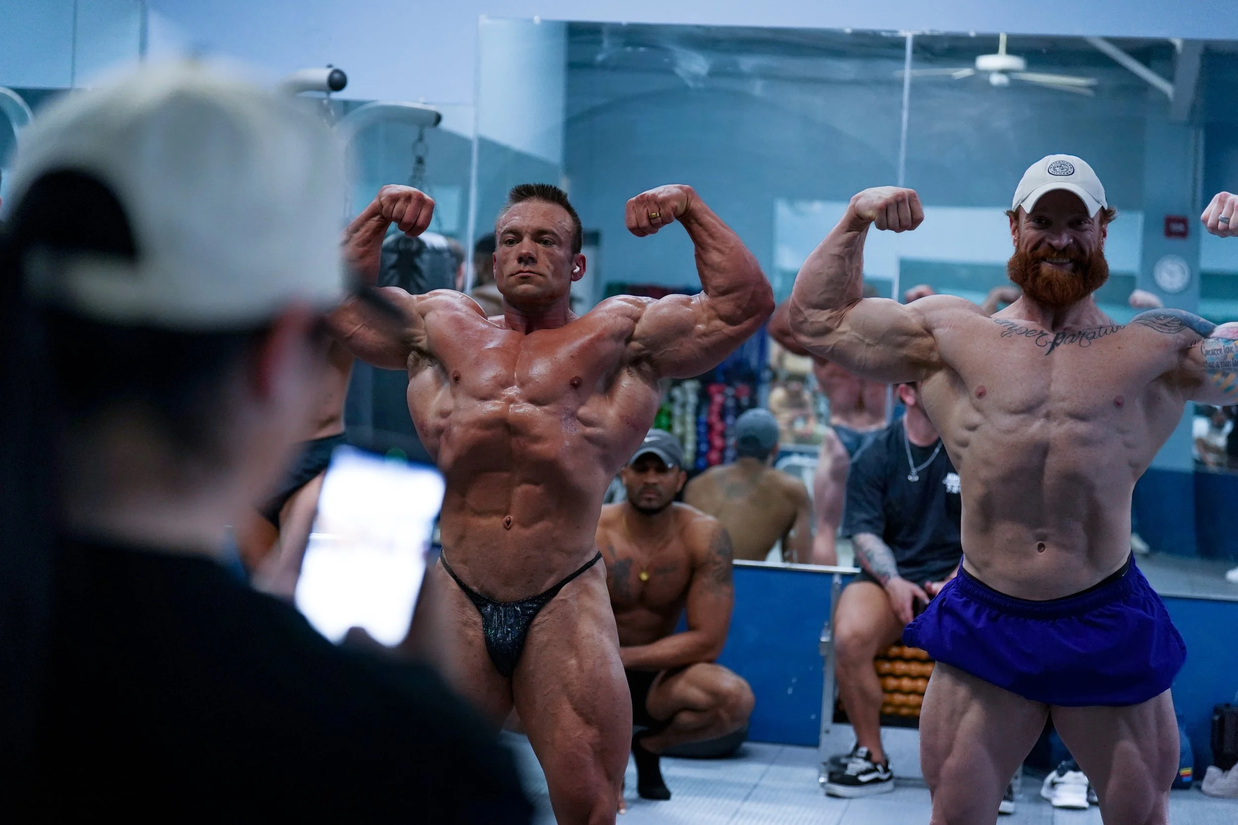 Two muscular men flexing their biceps in front of a gym mirror, with other people in the background.