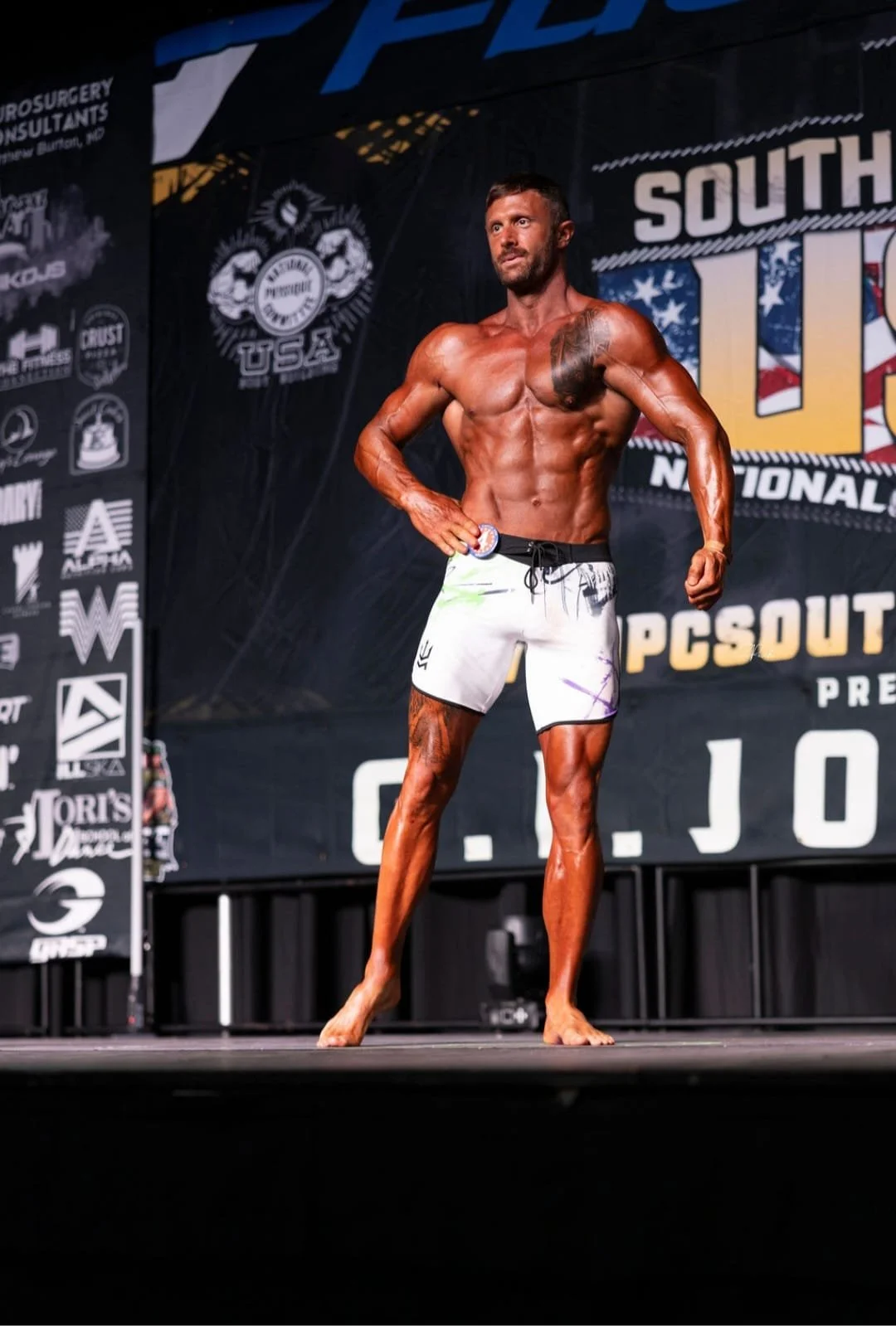 Male bodybuilder posing on stage during a competition, wearing white shorts, with elaborate muscular definition and tattoos, with a dark background featuring various sponsor logos.