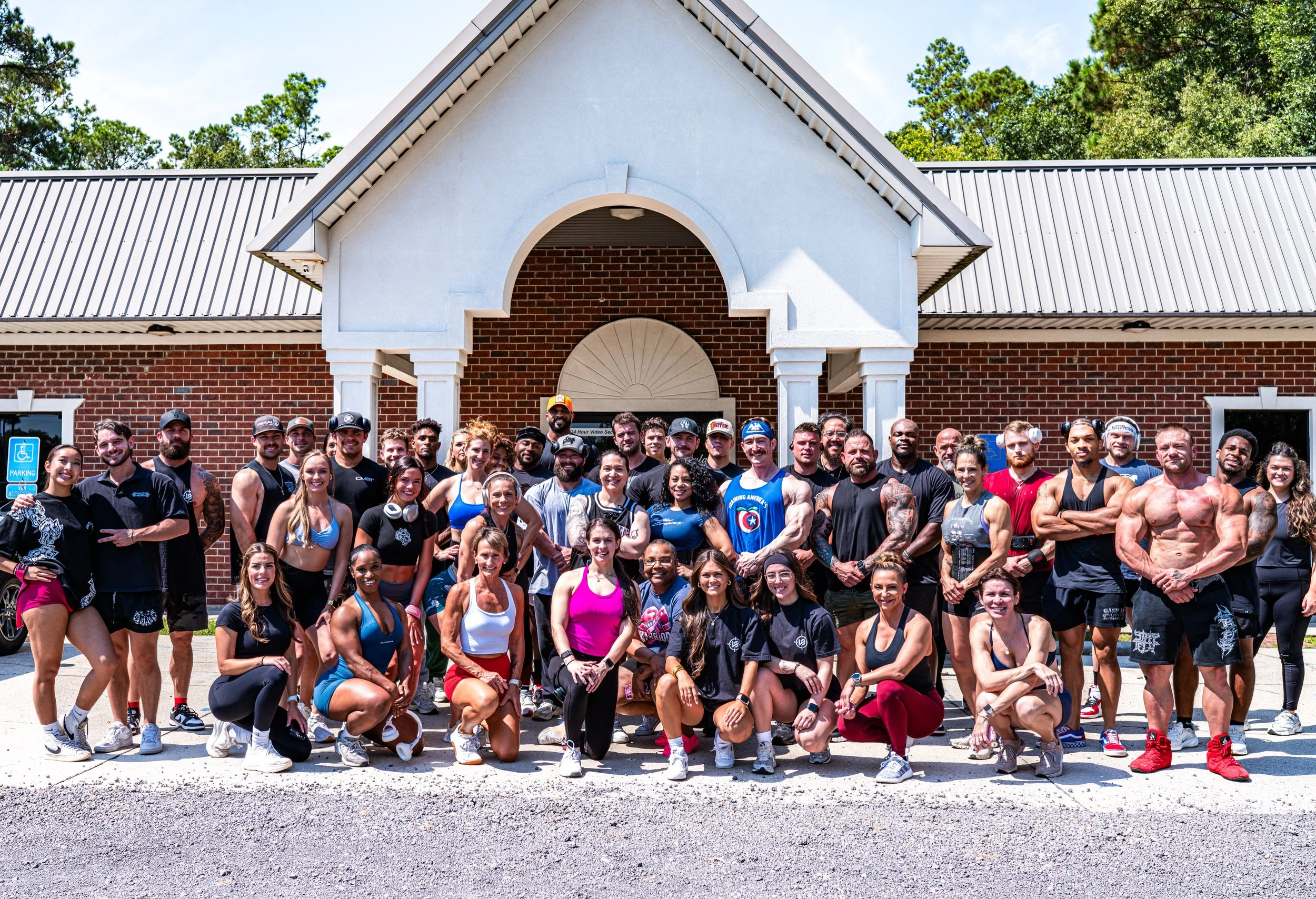 A diverse group of people posing outside a brick building with a white arch and metal roof, under a blue sky with green trees in the background.