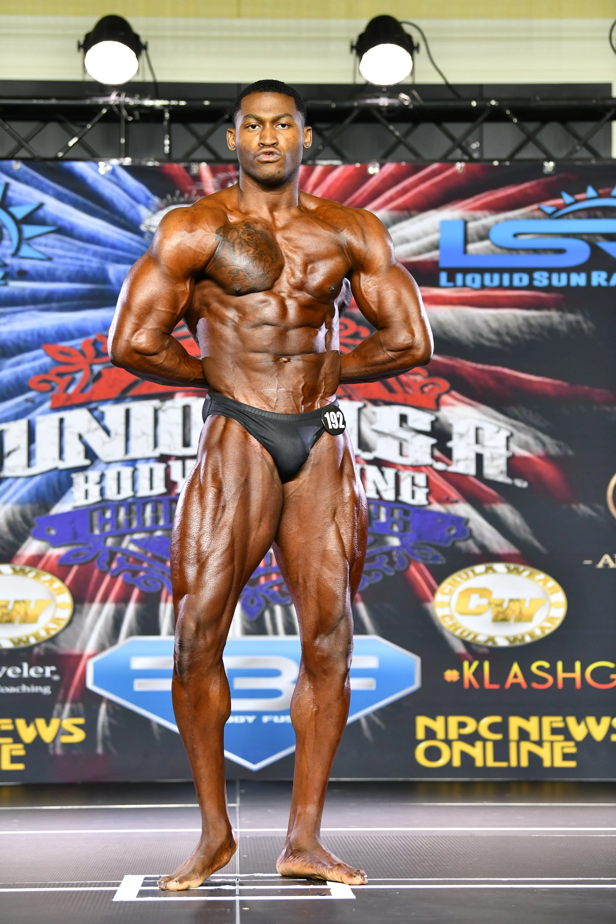 A male bodybuilder with dark skin wearing a black posing suit and standing on stage during a bodybuilding competition. He is flexing his muscles and posing in front of a backdrop with event logos.