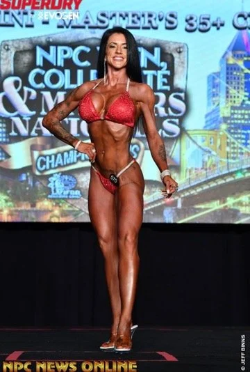 Female bodybuilder posing onstage during a competition, wearing a red bikini. The background displays a competition banner with graphics and text. Stage lights highlight her muscular physique as she stands in a confident stance, smiling.