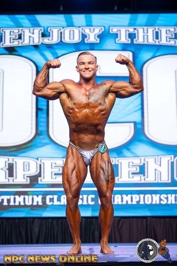 Bodybuilder posing on stage at a competition with "Enjoy the NPC Experience" banner in the background.