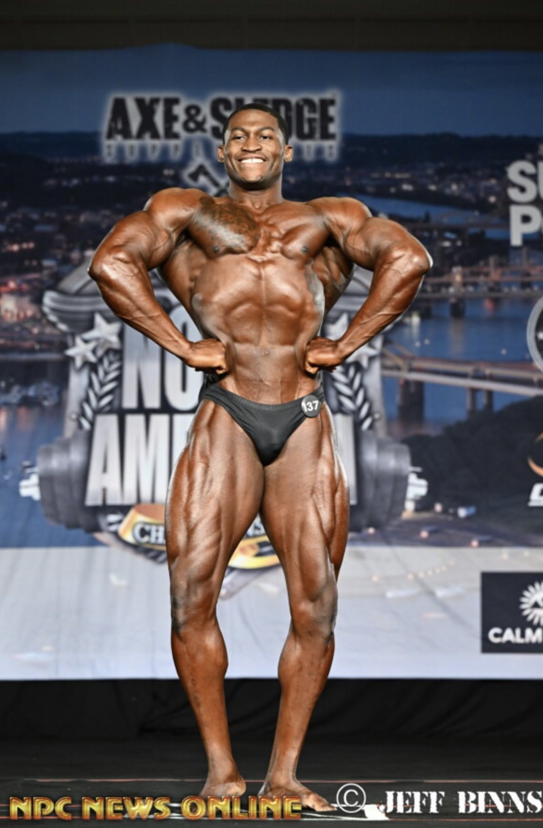 Male bodybuilder posing on stage, wearing black posing trunks, with a muscular physique and tanned skin, at a bodybuilding competition.