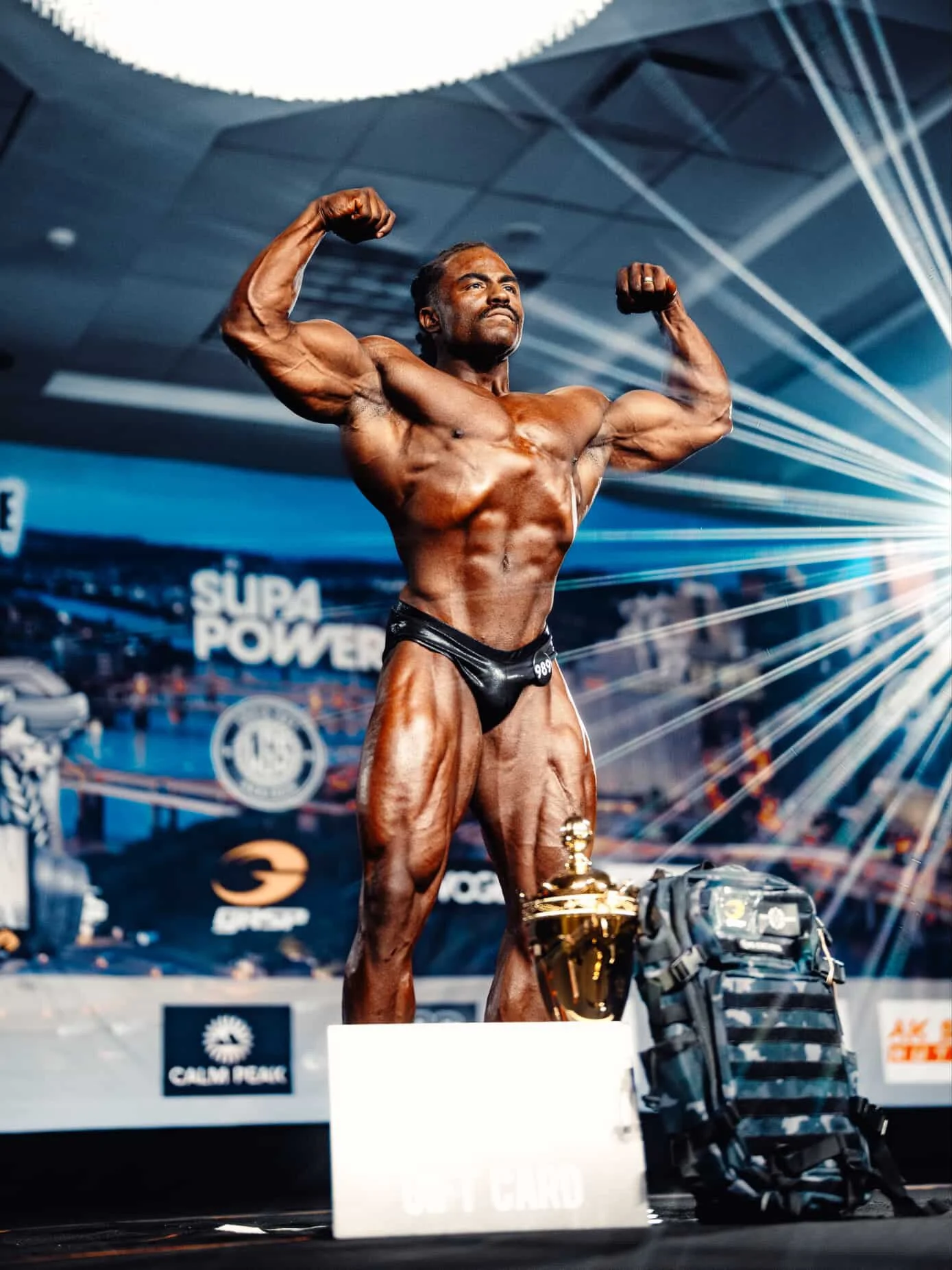 Male bodybuilder on stage in competition pose with flexed muscles, trophies, and a backpack nearby, in a well-lit arena with banners and audience in background.