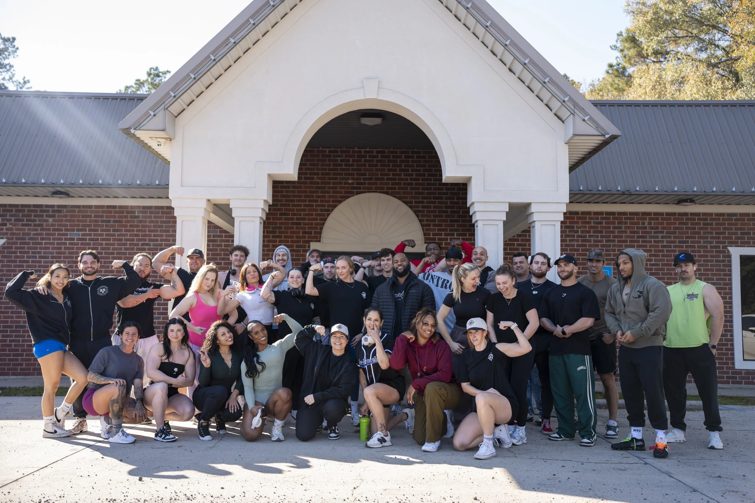 Group of diverse young adults outdoors in front of a brick building, posing with flexed arms and smiling.